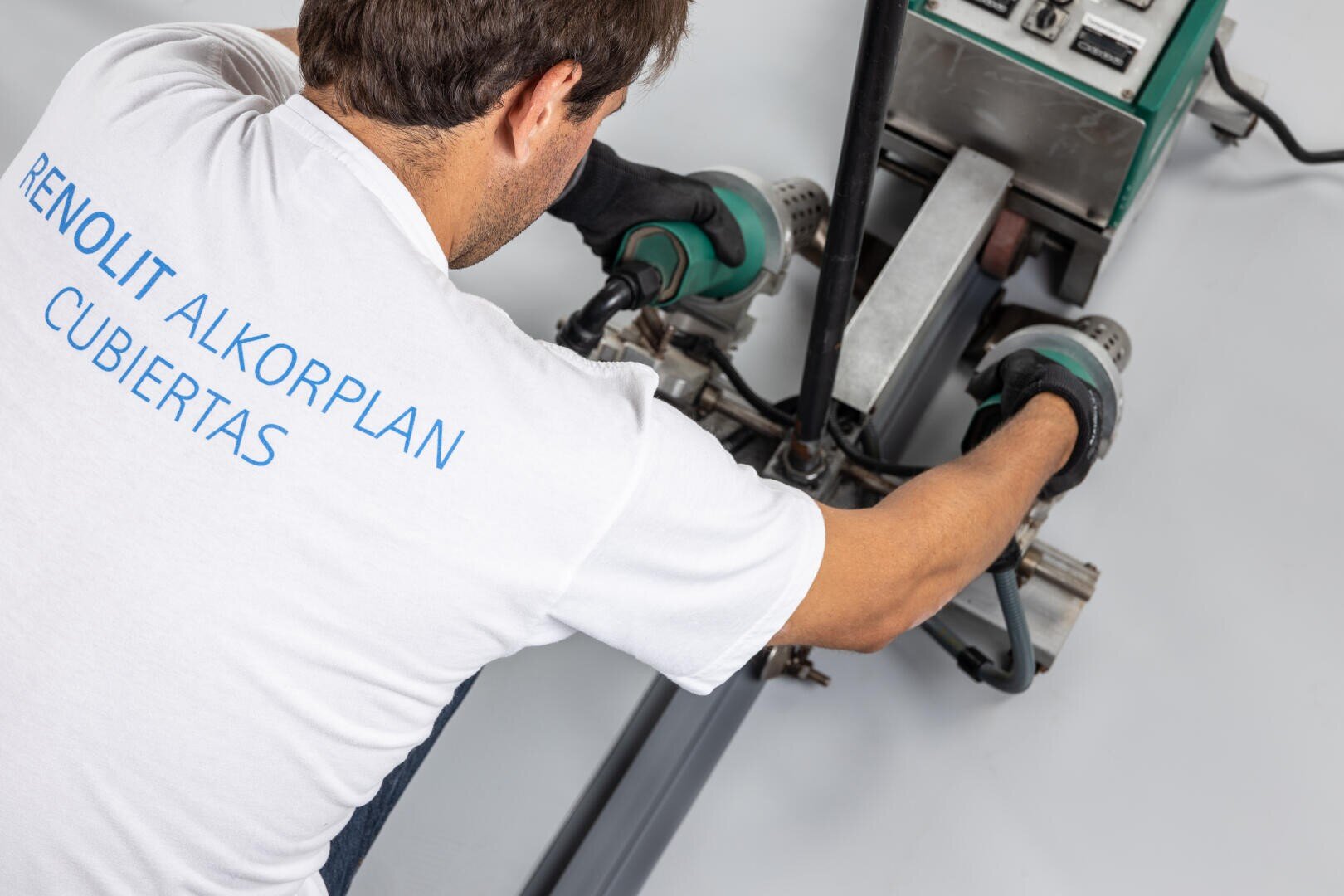 A worker wearing a white shirt with the inscription "RENOLIT ALKORPLAN CUBIERTAS" operates a green industrial machine and presses it along a seam onto a flat, gray surface.