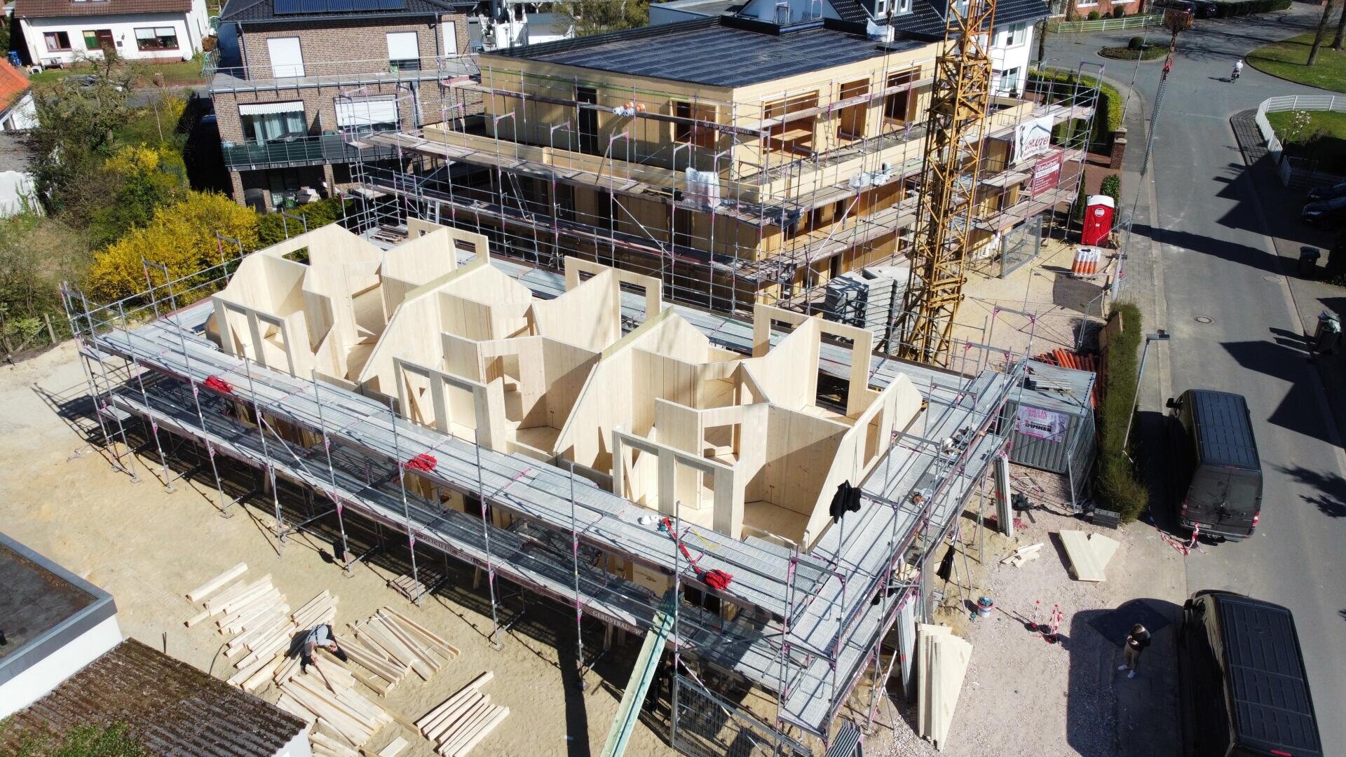 Aerial view of a construction site with a wooden panel building under construction. The structure is surrounded by scaffolding and workers, materials and equipment can be seen. An adjacent building appears to be still under construction.