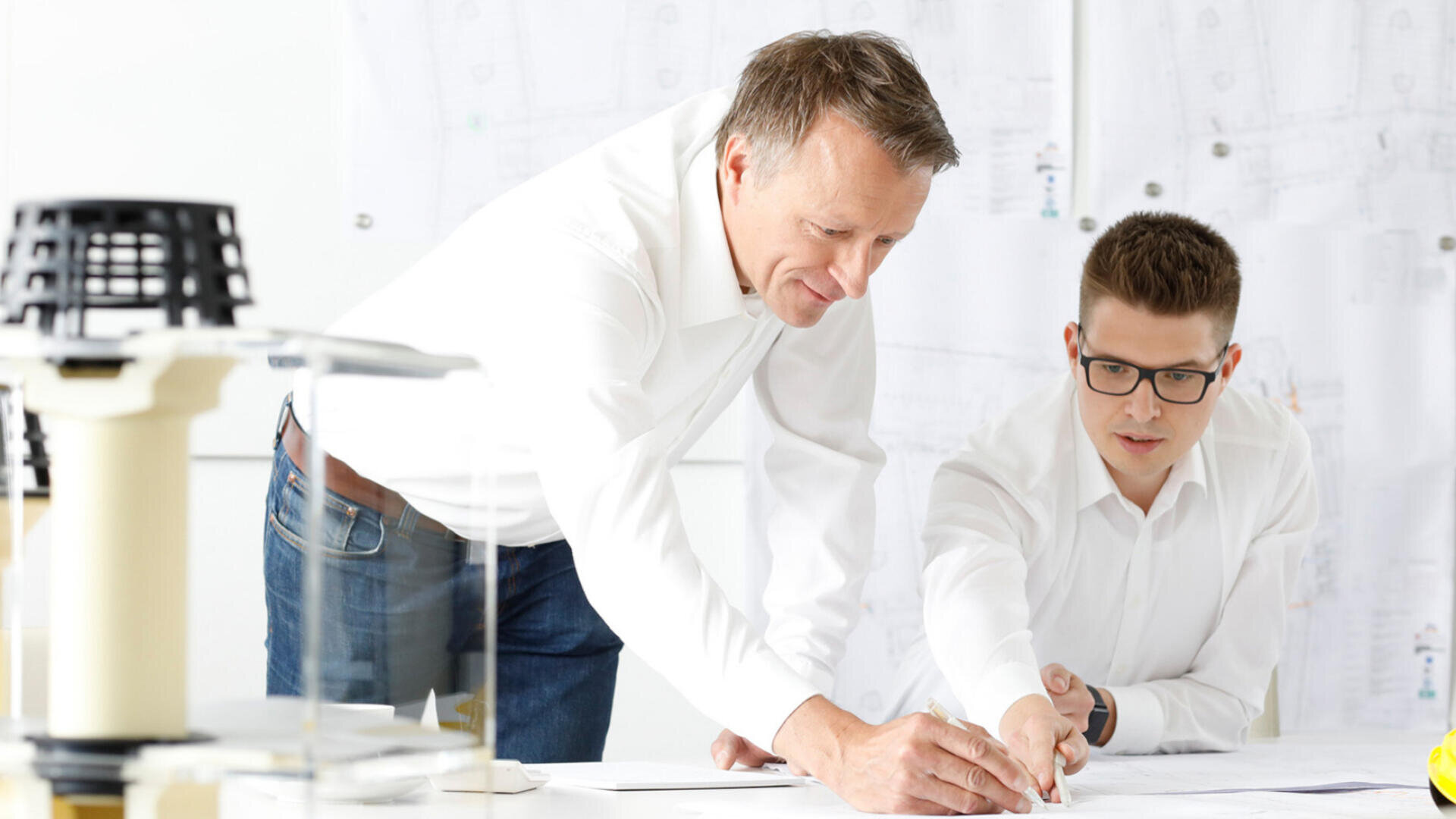 Zwei Männer in weißen Hemden arbeiten gemeinsam an einem Schreibtisch, der mit Papieren und technischen Zeichnungen bedeckt ist. Einer der Männer beugt sich vor und zeigt auf etwas auf den Papieren, während der andere konzentriert und engagiert zuzuschauen scheint.