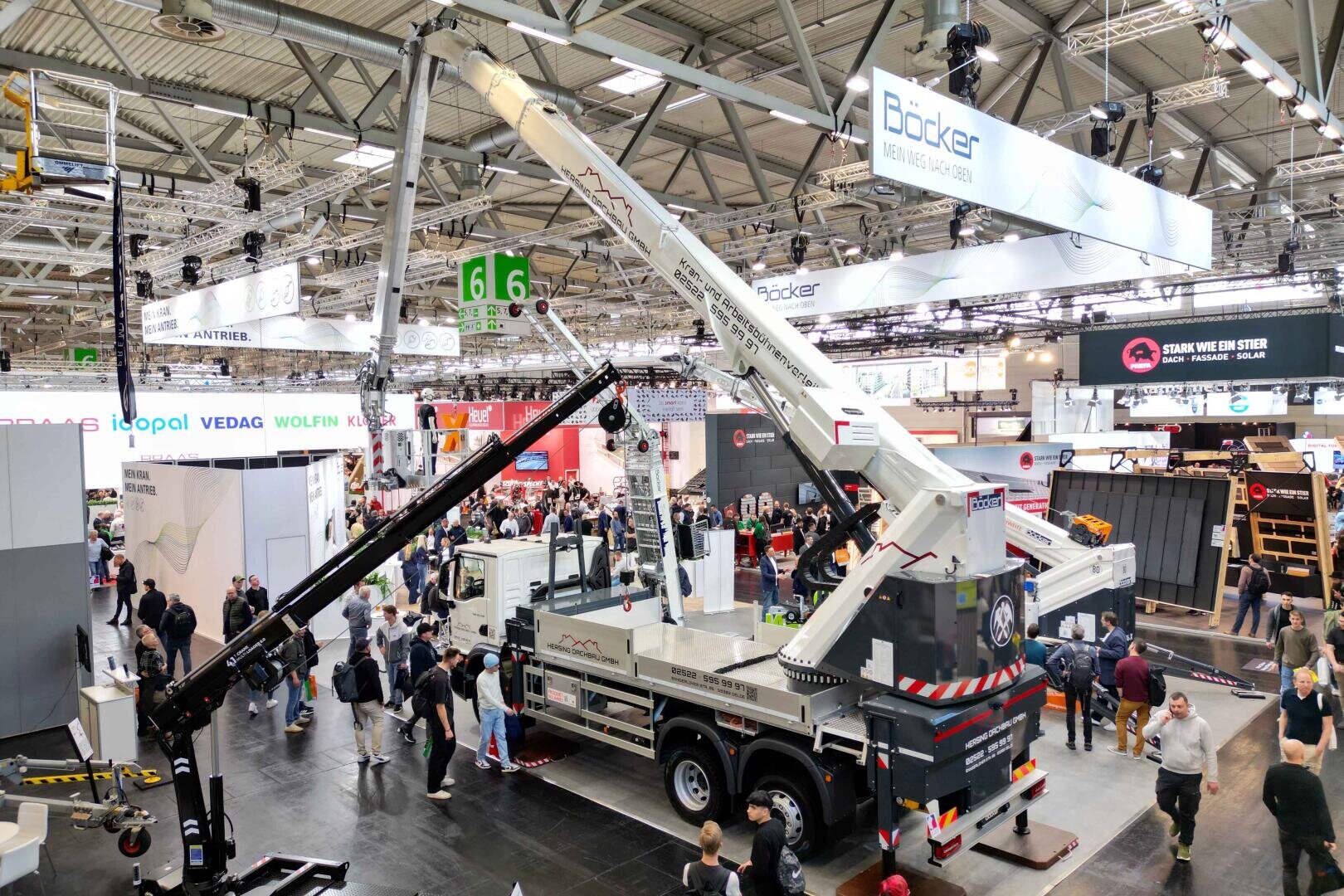 In einer belebten Ausstellungshalle steht ein großer weißer Kranwagen, der von Besuchern umringt ist. Überkopfbanner und Stände von verschiedenen Unternehmen füllen den hell erleuchteten Raum.