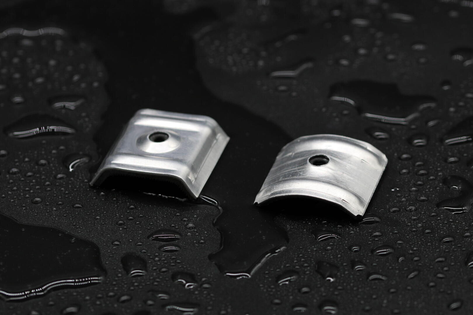 Zwei kleine, gebogene Metallklammern mit einem Loch in der Mitte werden auf eine nasse, schwarze Oberfläche gestellt, auf der Wassertropfen verstreut sind.