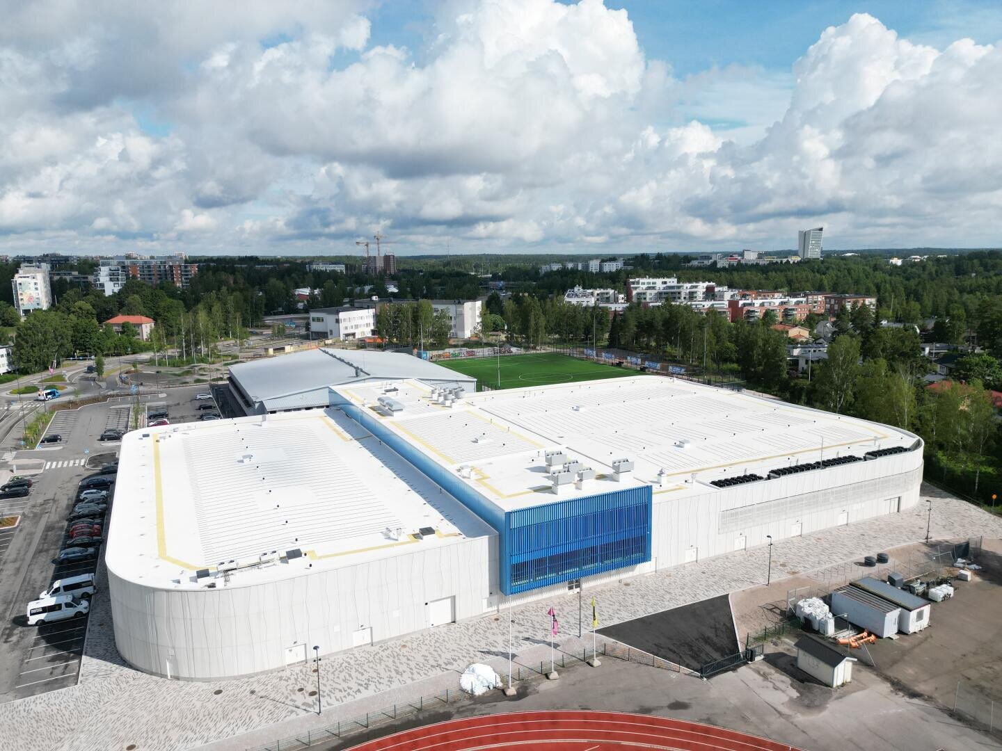 Ein großes, modernes weißes Gebäude mit Flachdach und blauen Akzenten steht neben einem Sportplatz und einer Laufbahn, umgeben von Bäumen und Wohngebäuden unter einem bewölkten Himmel.
