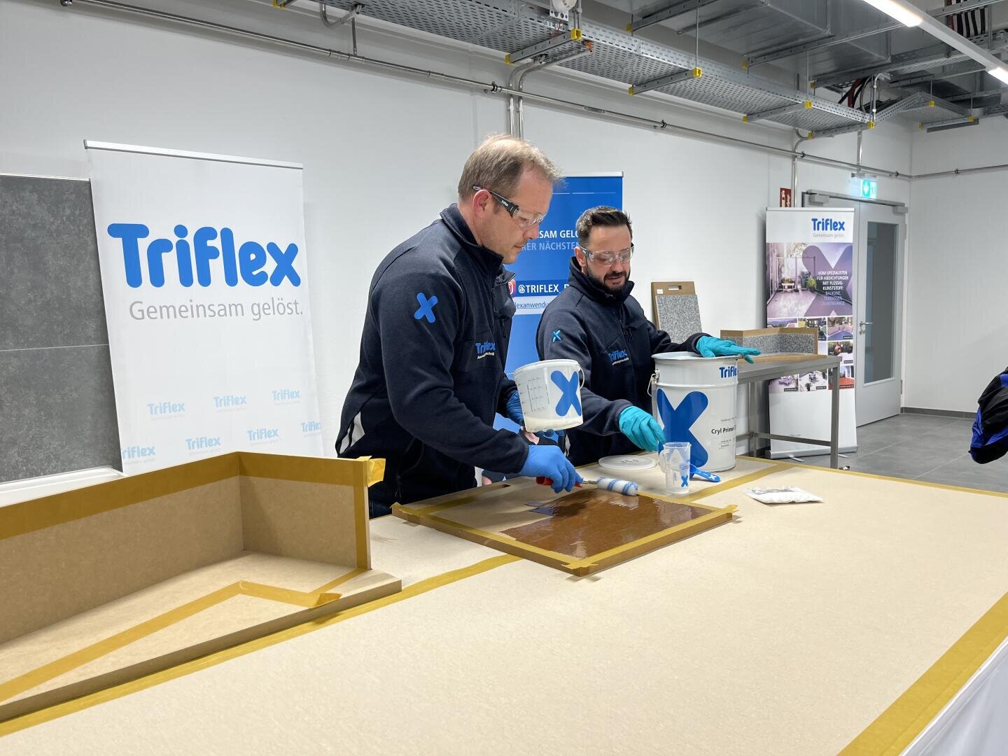 Two men in Triflex uniforms apply a liquid substance from buckets onto a surface in a modern workshop. Triflex branding and promotional materials are visible in the background.