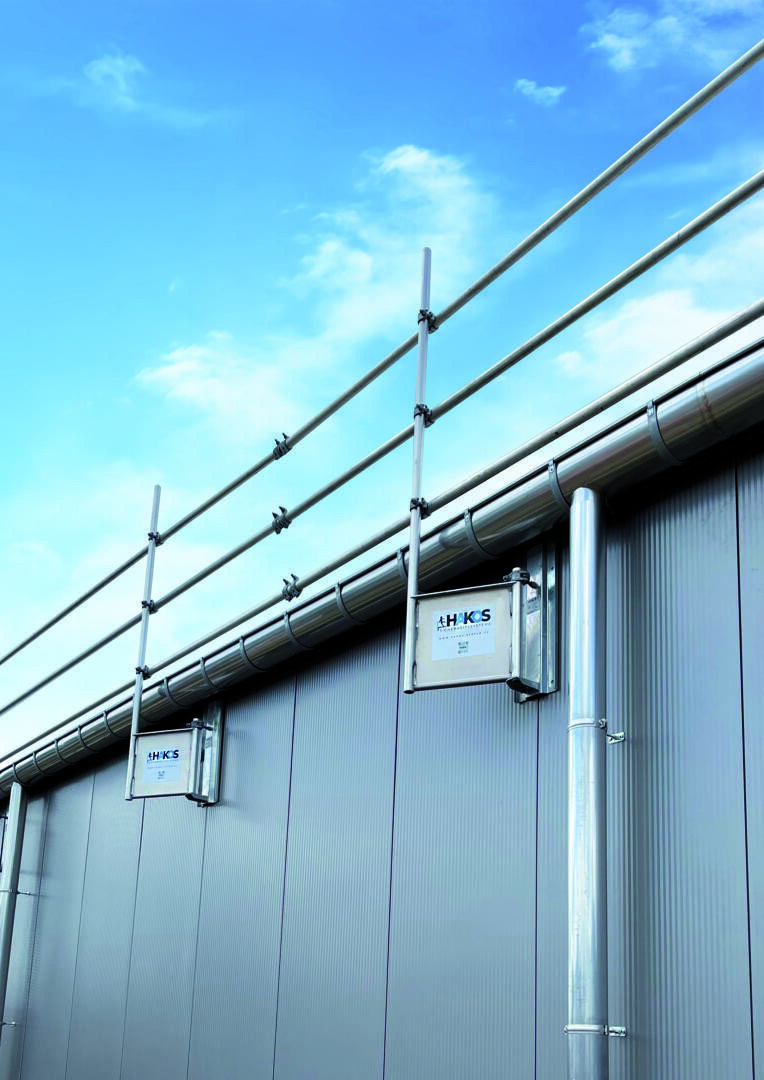 Metal railing on the edge of the roof of a modern industrial building, with blue sky and some clouds in the background. The building has a corrugated iron exterior and visible signage.