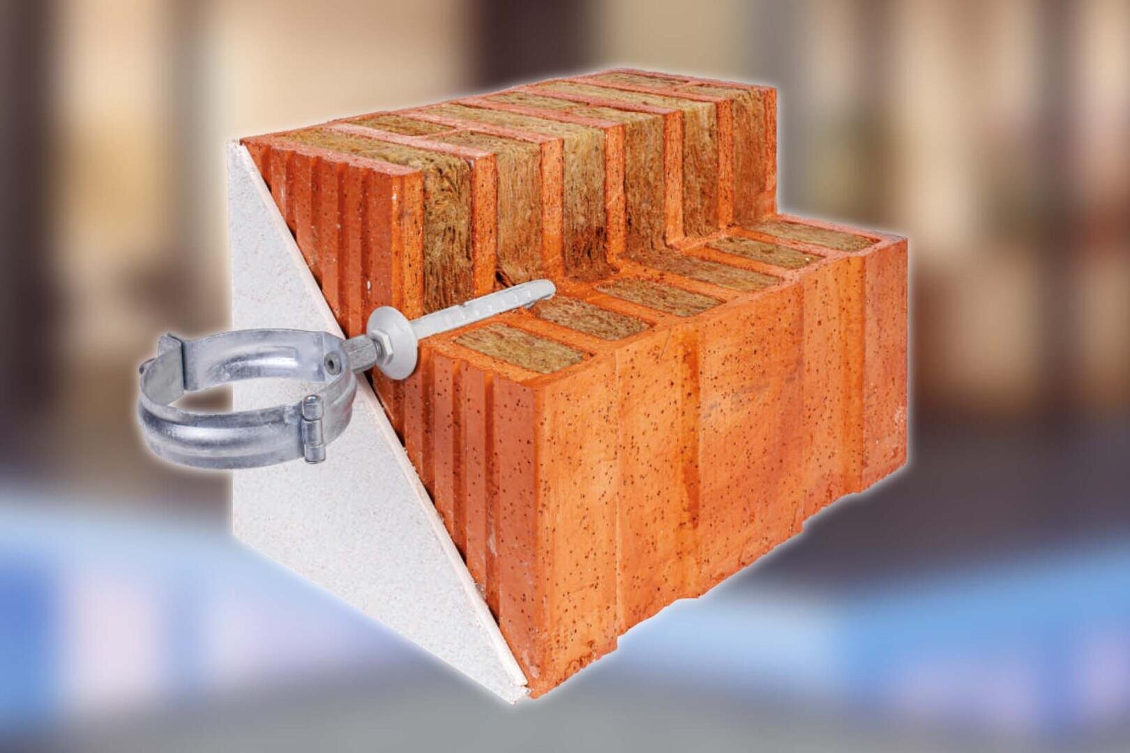 Eine Nahaufnahme eines aufgeschnittenen roten Ziegelsteins mit horizontalen Rillen, die seine innere Struktur zeigen, und einer Metallklammer, die mit einer Schraube an der linken Seite befestigt ist. Der Hintergrund ist unscharf.