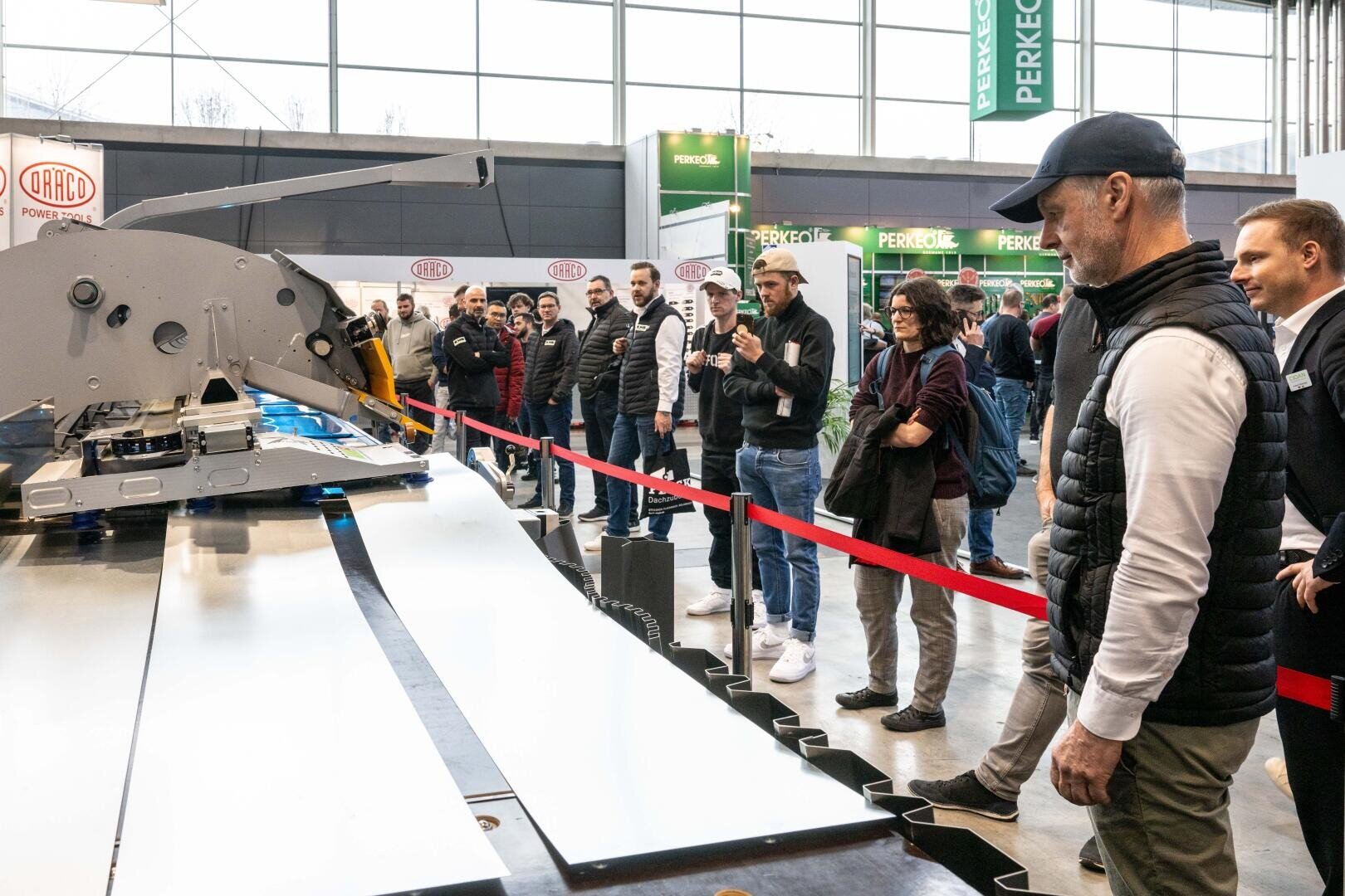 Eine Gruppe von Personen beobachtet eine große industrielle Schneidemaschine in einer Indoor-Ausstellung in Aktion. Die Szene ist durch ein rotes Seil abgetrennt, und die Besucher beobachten und fotografieren.