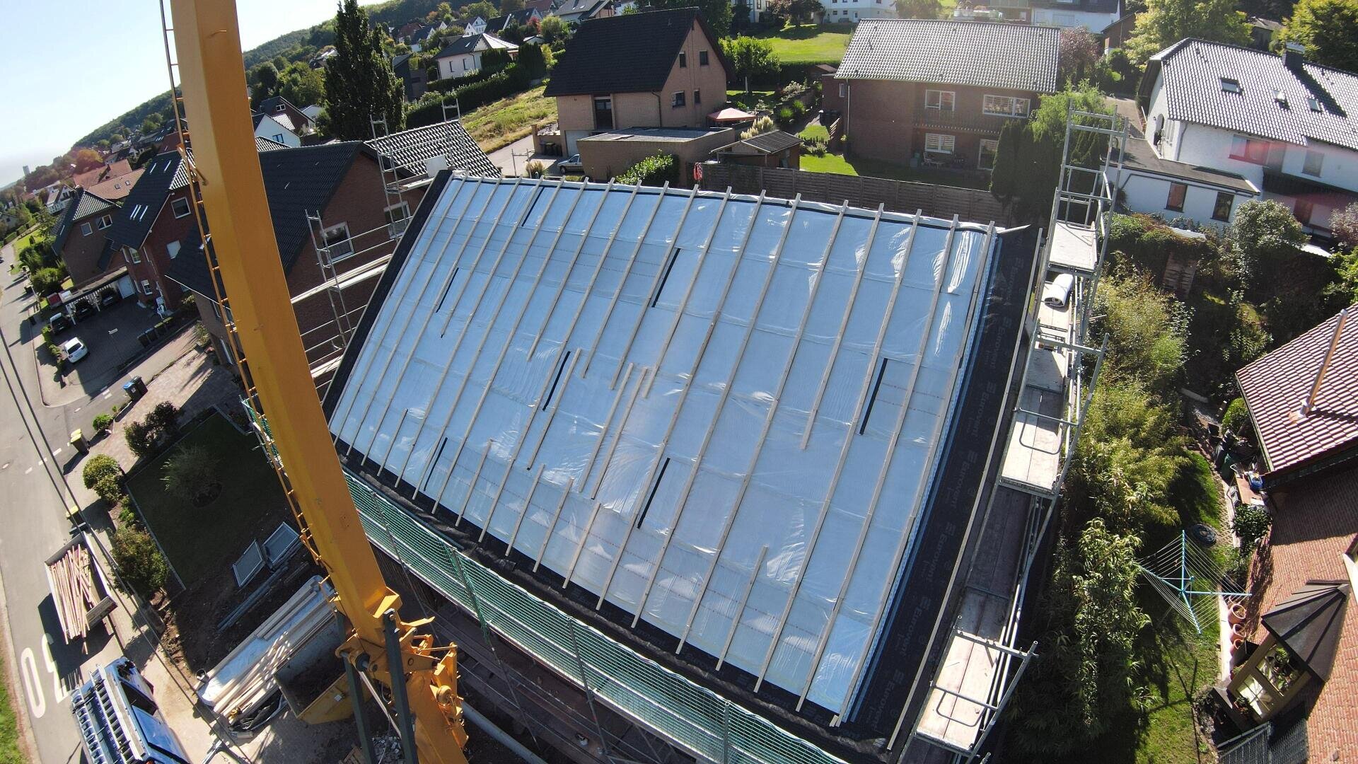 Aerial view of a house roof under construction, covered with a reflective silver material and surrounded by scaffolding in a residential area. A crane can be seen on the left side of the picture.