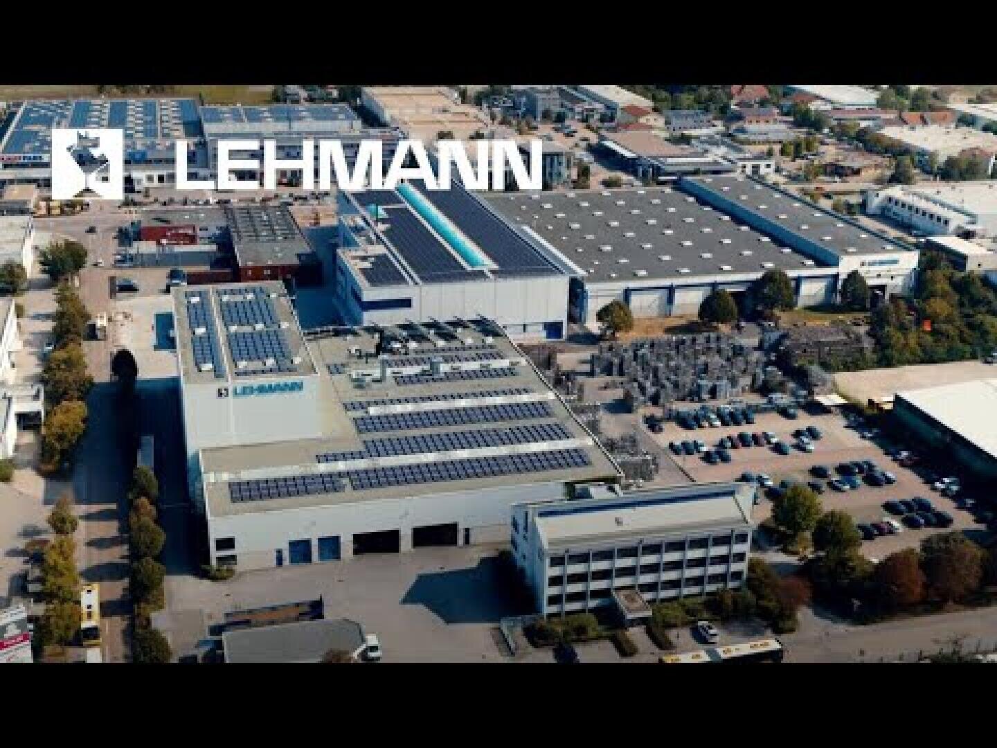 Aerial view of the LEHMANN industrial site with several large buildings with solar cells on the roofs, parking lots and surrounding green areas. The LEHMANN logo is clearly visible in the upper part of the picture.