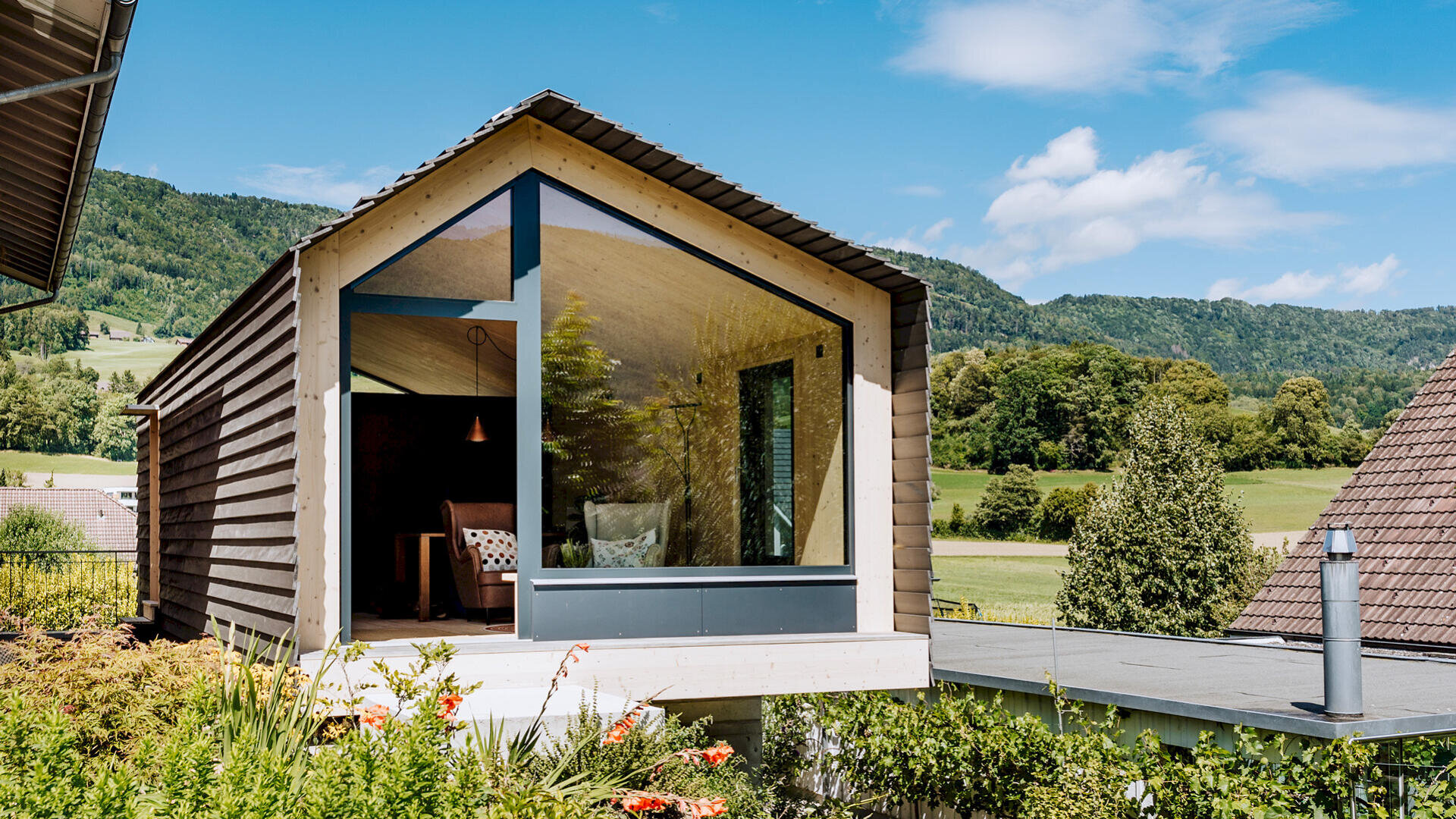 Modernes Haus mit großem dreieckigem Glasfenster und Holzverkleidung, umgeben von Grün und Hügeln unter blauem Himmel mit vereinzelten Wolken. Im Inneren befinden sich ein Sofa und eine Hängelampe, die durch das Fenster sichtbar sind.