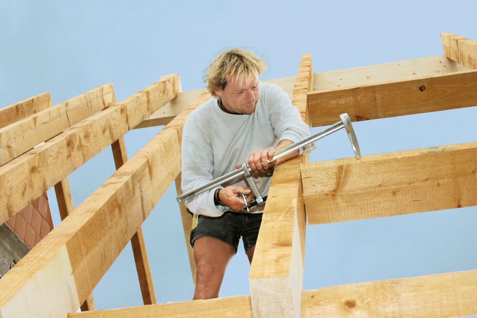 Eine Person, die ein hellgraues Sweatshirt und Shorts trägt, verwendet ein Werkzeug, um eine Holzrahmenkonstruktion unter einem klaren blauen Himmel zusammenzusetzen.