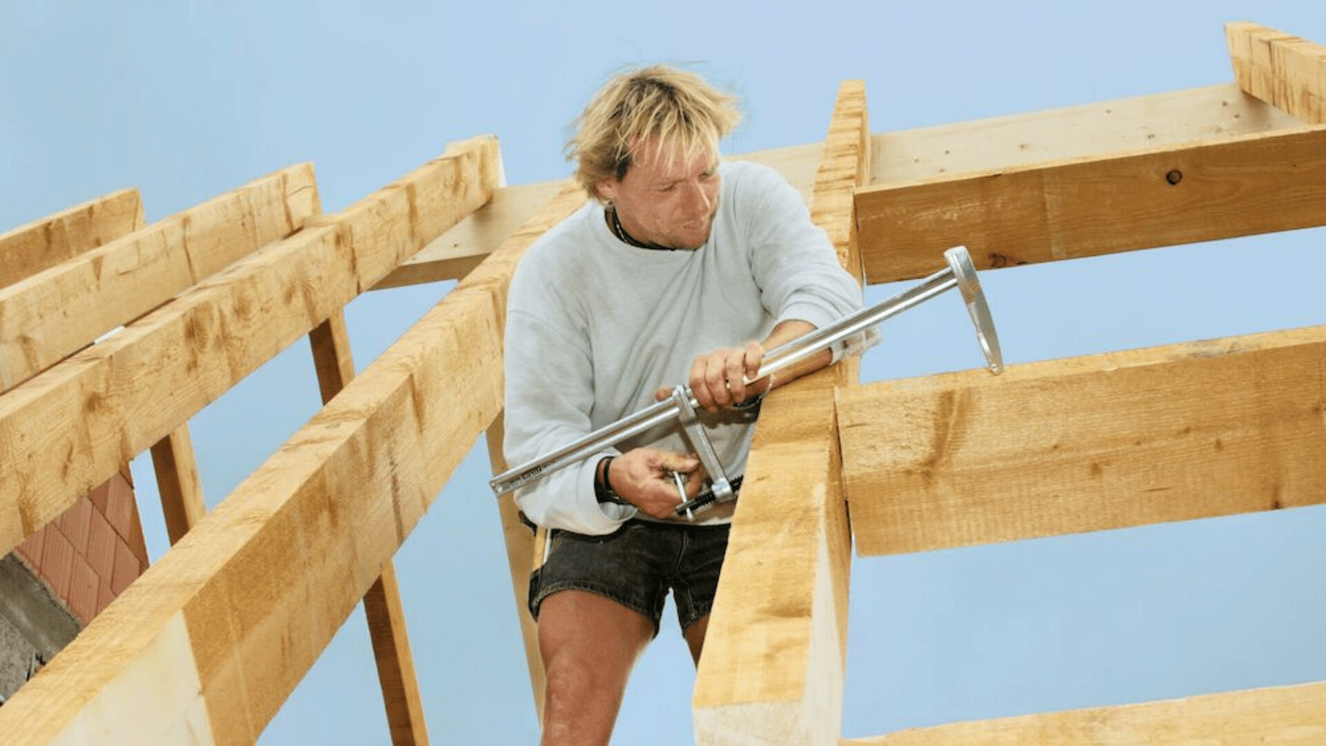 Eine Person in hellem Hemd und Shorts arbeitet vor blauem Himmel mit einem Werkzeug an einem hölzernen Dachstuhl.