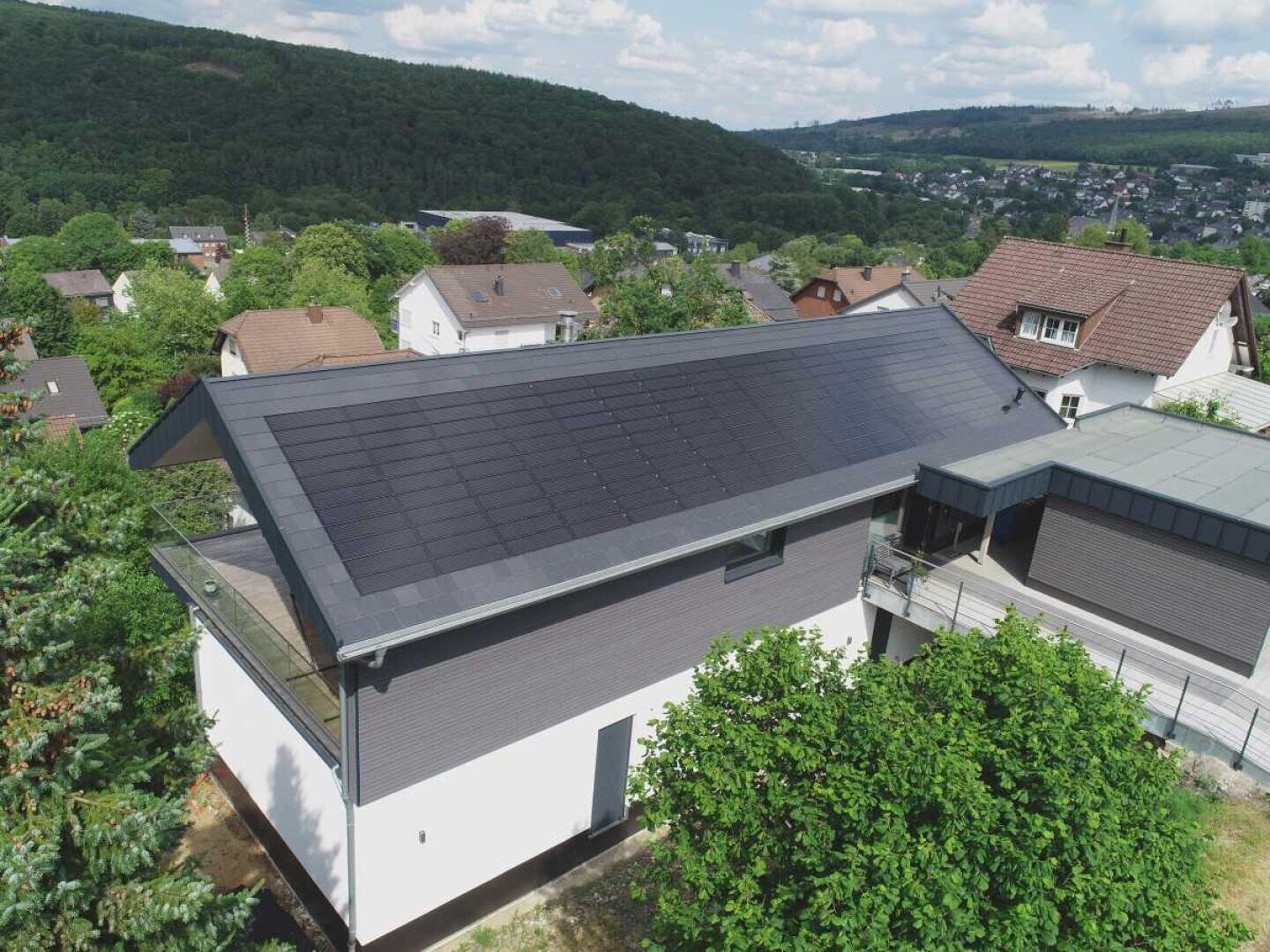Ein modernes Haus mit einer großen Reihe von Sonnenkollektoren auf dem Dach, umgeben von Bäumen und Nachbarhäusern in einer hügeligen, grünen Vorstadtgegend unter einem teilweise bewölkten Himmel.