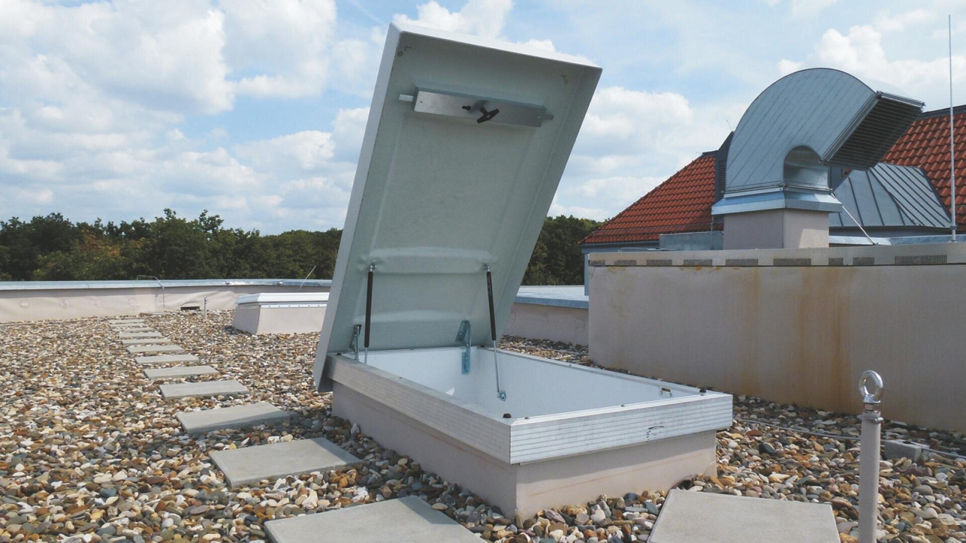 Auf einem mit Kies bedeckten Flachdach ist eine Metalldachluke geöffnet, zu der Trittsteine aus Beton führen. In der Nähe befinden sich eine große Lüftungsanlage und ein Gebäude mit einem roten Ziegeldach im Hintergrund.