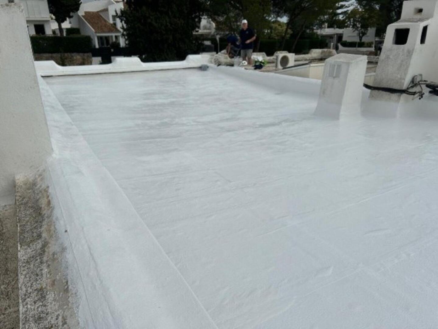 Ein flaches weißes Dach mit frischem Anstrich, auf dem im Hintergrund zwei Personen in der Nähe von Werkzeugen und Geräten arbeiten. Um das Dach herum sind Wohngebäude und Grünflächen zu sehen.