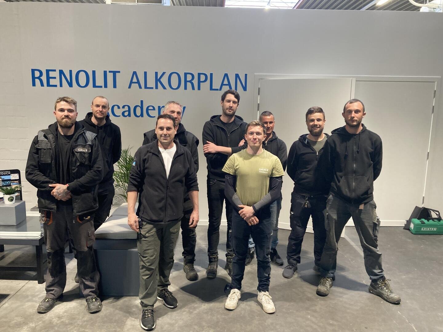 Nine men stand and pose for a group photo inside a building with white walls. The background displays a sign reading RENOLIT ALKORPLAN academy. They are dressed in casual work attire.
