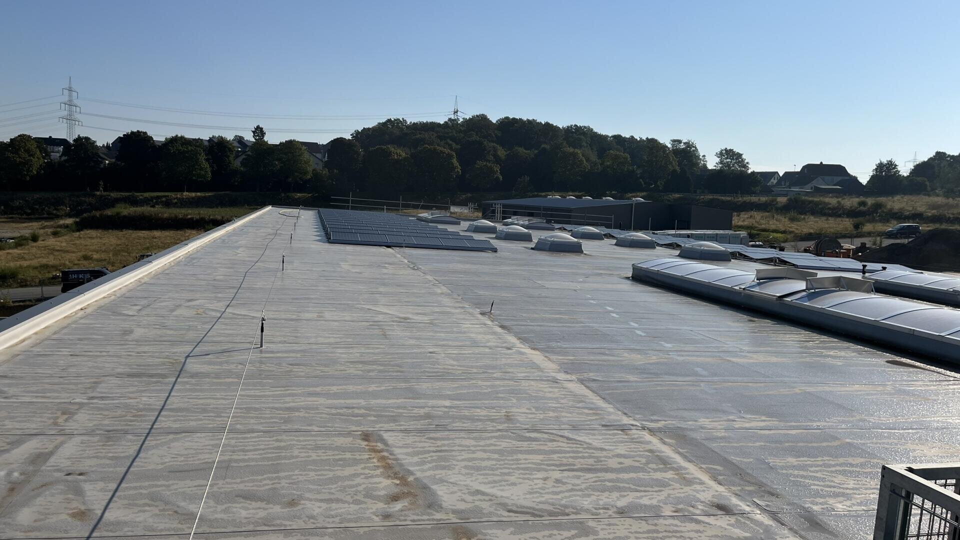 A large, flat industrial rooftop with solar panels and dome-shaped skylights, photographed on a sunny day. Trees, grassy hills, utility poles, and a few buildings are visible in the background.