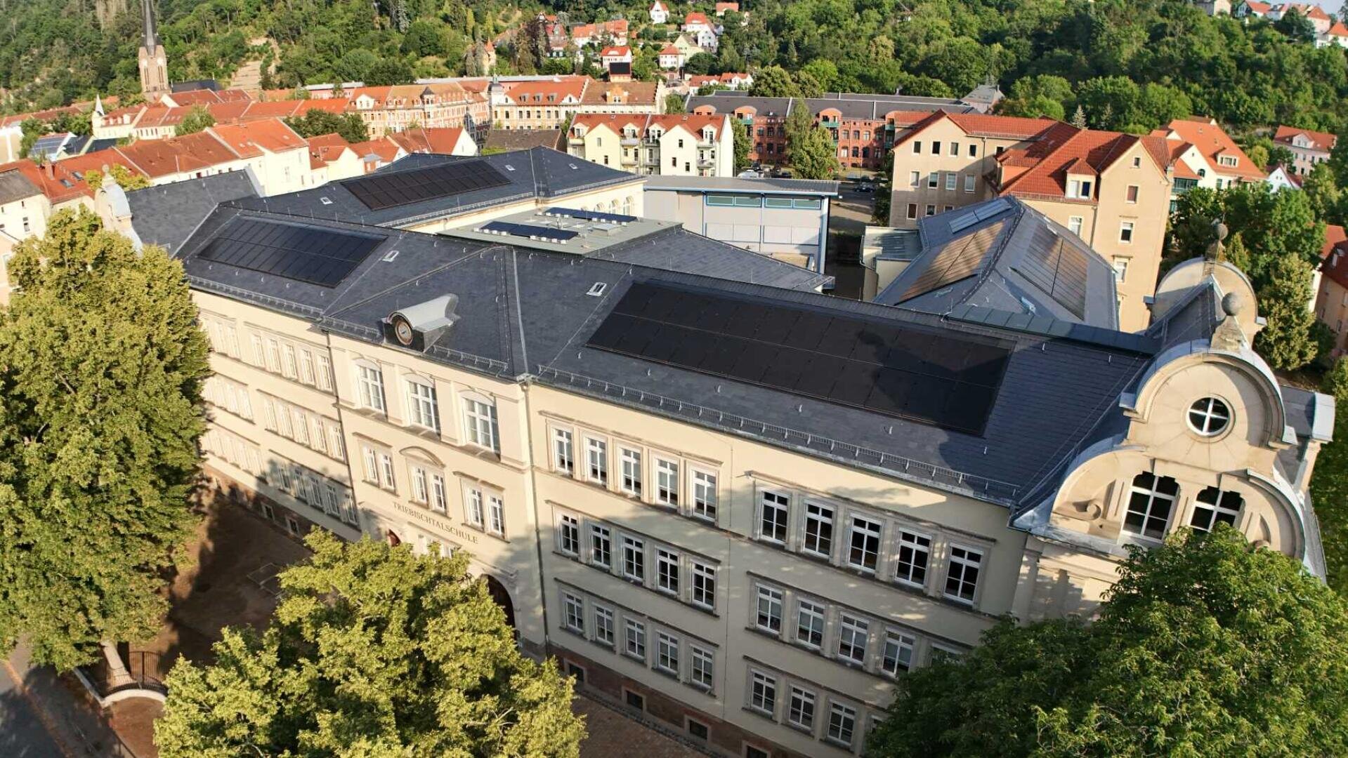 Luftaufnahme eines großen, hellen Gebäudes mit Sonnenkollektoren auf dem Dach, umgeben von Bäumen und anderen Gebäuden in einer grünen, hügeligen Gegend.