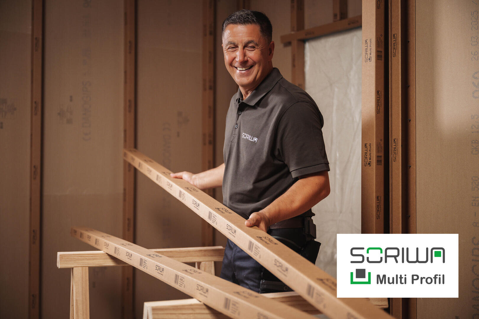 A smiling man in a SORIWA polo shirt holds two wooden planks in a room under construction. The SORIWA Multi Profile logo appears in the bottom right corner.