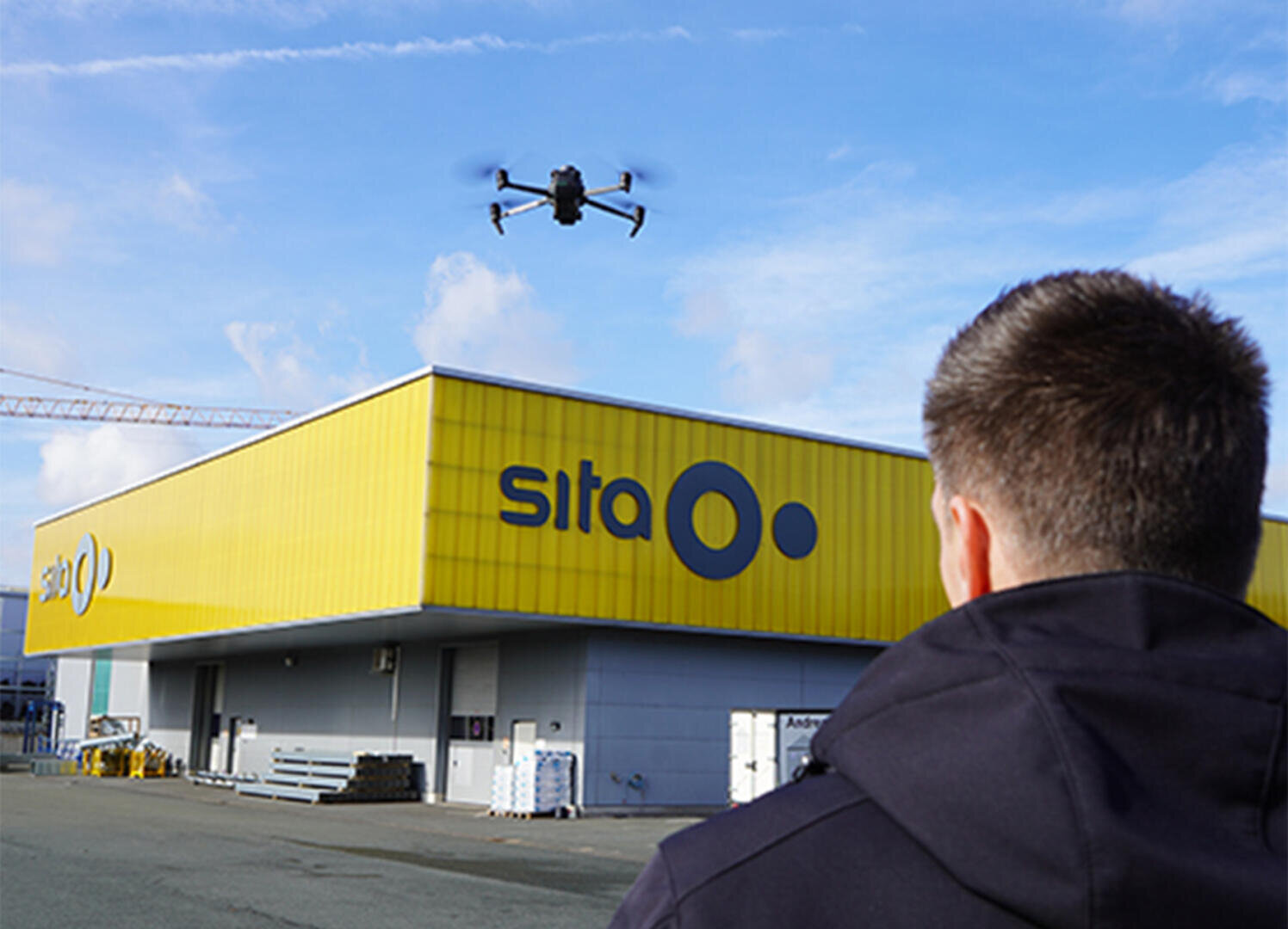 Eine Person in einer dunklen Jacke steuert eine fliegende Drohne vor einem großen gelben Gebäude mit der Aufschrift sita unter einem teilweise bewölkten Himmel.