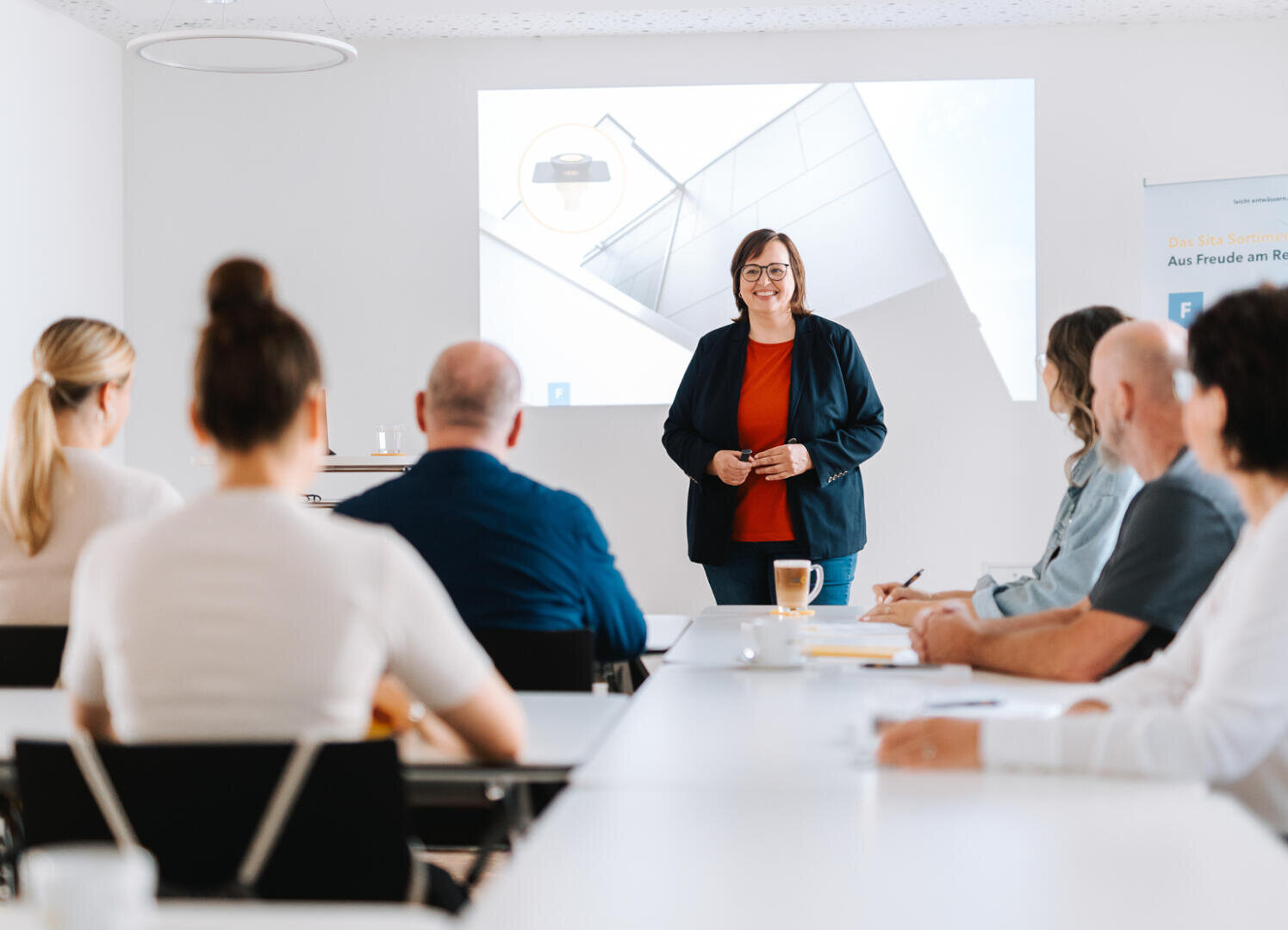 Eine Frau steht und lächelt, während sie einer Gruppe von Personen, die an Tischen in einem hellen, modernen Konferenzraum sitzen, eine Präsentation hält. Hinter ihr ist eine Projektion zu sehen.