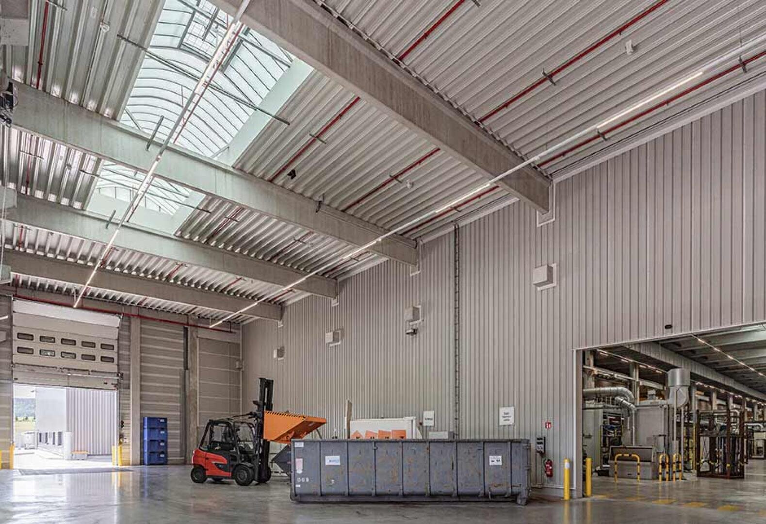 Ein geräumiges Industrielager mit hohen Decken, Oberlichtern, einem Gabelstapler, der Material transportiert, einem großen grauen Müllcontainer und sichtbaren Rohren an einer Wand. Der Bereich erscheint sauber und gut beleuchtet.