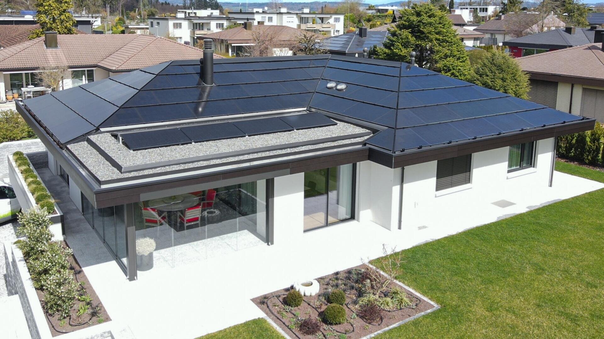 Modernes einstöckiges Haus mit dunklen Solardachziegeln, großen Fenstern, einer Terrasse mit Sitzgelegenheiten im Freien und einem gepflegten Rasen und Garten, umgeben von anderen Vorstadthäusern und Grünanlagen.