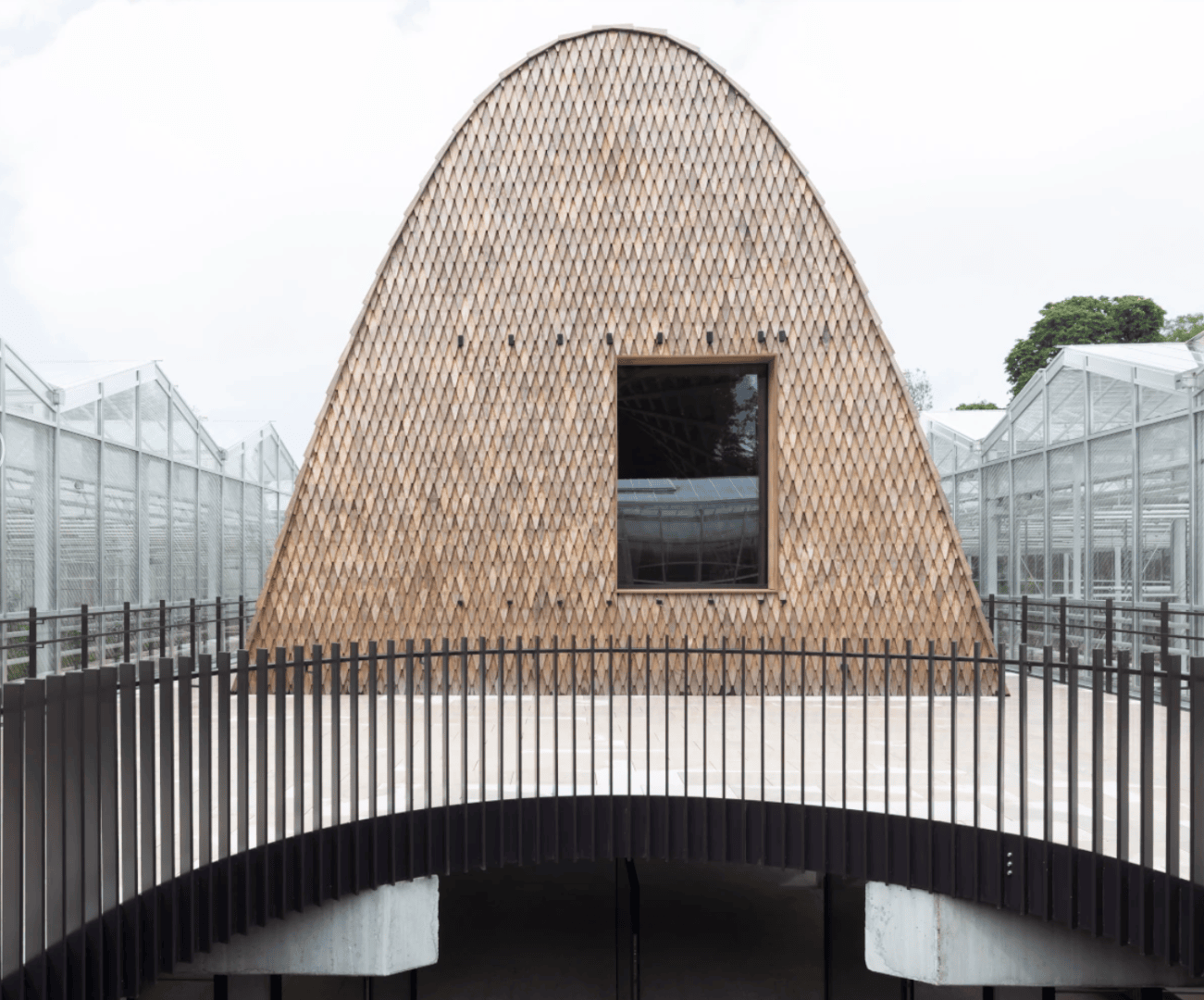 Ein kuppelförmiges Gebäude mit einer strukturierten, gewebten Außenseite und einem einzigen quadratischen Fenster. Ein geschwungenes schwarzes Metallgeländer führt zum Eingang hinauf. Im Hintergrund sind Glasstrukturen und Grünflächen zu sehen.
