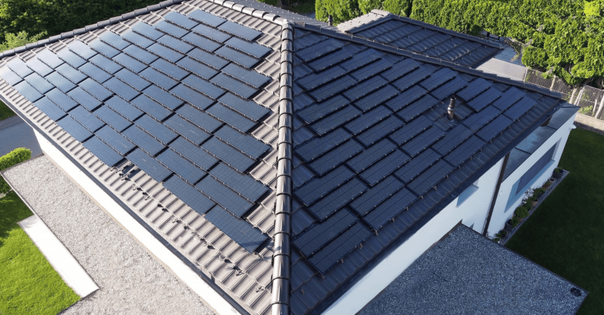 Ein mit dunklen Solarziegeln gedecktes Hausdach, das sich nahtlos in die traditionellen Dachziegel einfügt, umgeben von grünem Gras und Bäumen. Die Solarziegel fangen das Sonnenlicht für erneuerbare Energie ein.