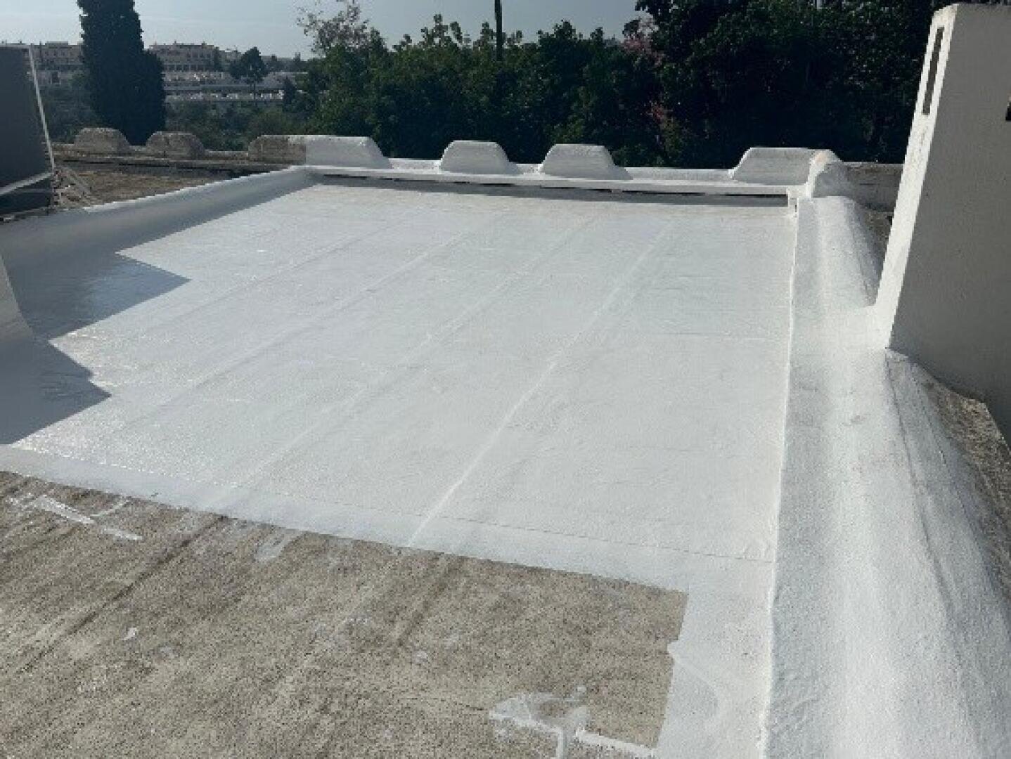 Ein flaches Dach mit einer frisch aufgetragenen weißen, wasserdichten Beschichtung, umgeben von niedrigen Brüstungsmauern und einigen erhöhten Dachelementen, unter einem klaren Himmel mit Bäumen und Gebäuden im Hintergrund.