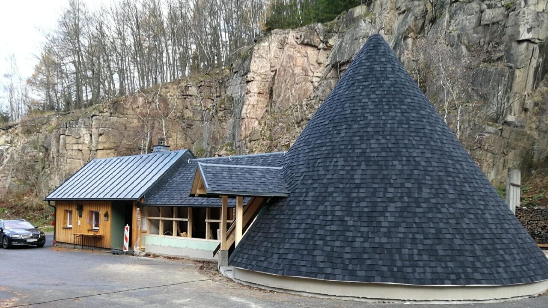 Ein einzigartiges Gebäude mit einem hohen, kegelförmigen Dach neben einem kleineren, rechteckigen Teil, vor einer felsigen Klippe mit kahlen Bäumen und einem geparkten Auto auf der linken Seite.