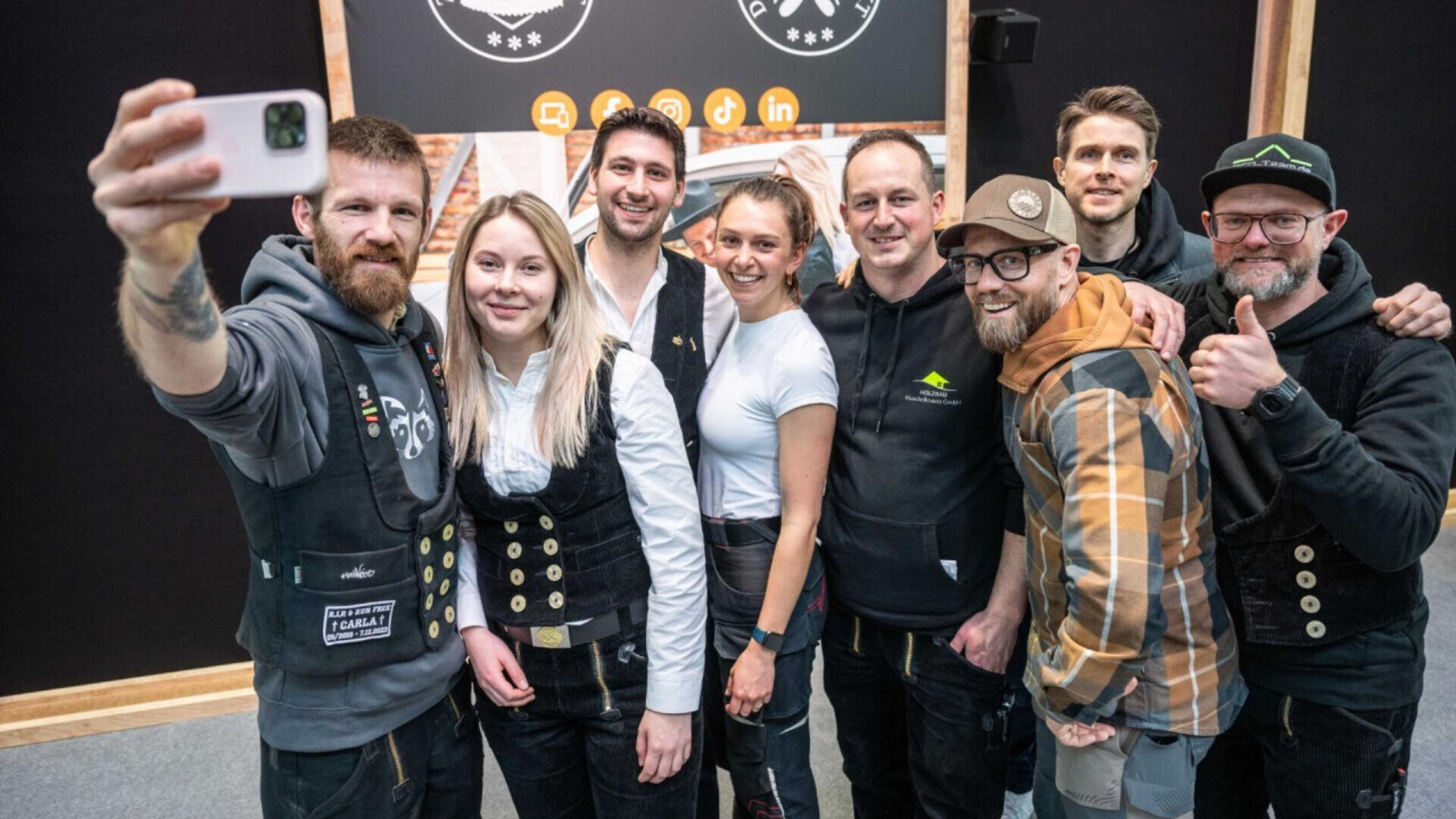A group of eight smiling people stand close together, posing for a selfie in front of a dark wall with logos. Most are wearing casual or work clothing, and one person in front holds up a smartphone to take the photo.