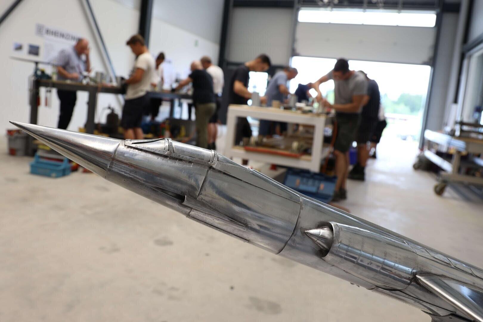 Eine Nahaufnahme eines metallischen Raketenmodells in einem Innenraum, mit mehreren Personen, die an Tischen im unscharfen Hintergrund an Projekten arbeiten. Die Umgebung scheint eine Werkstatt oder ein Labor zu sein.