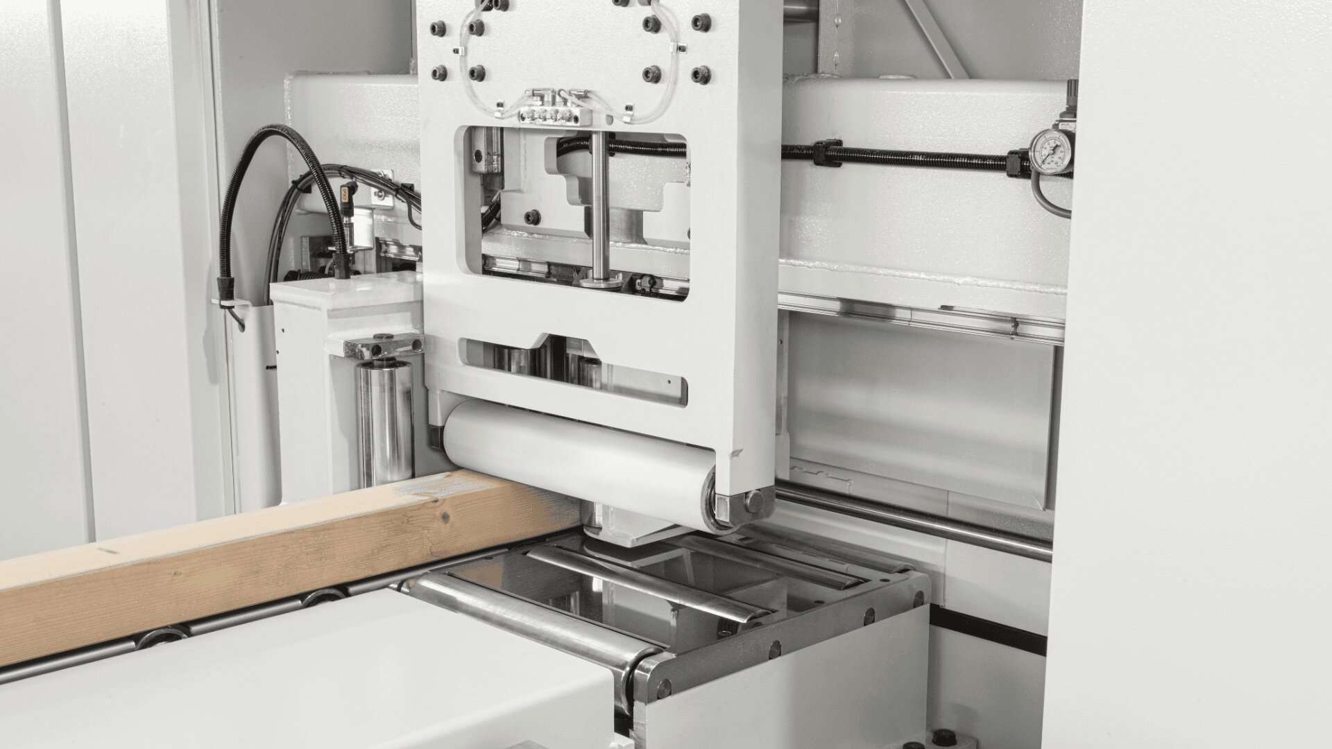 Nahaufnahme einer industriellen Holzverarbeitungsmaschine, die an einem Holzbalken arbeitet. Sie zeigt Metallkomponenten, Rollen und Hydraulikteile in einer weißen, sauberen Umgebung.