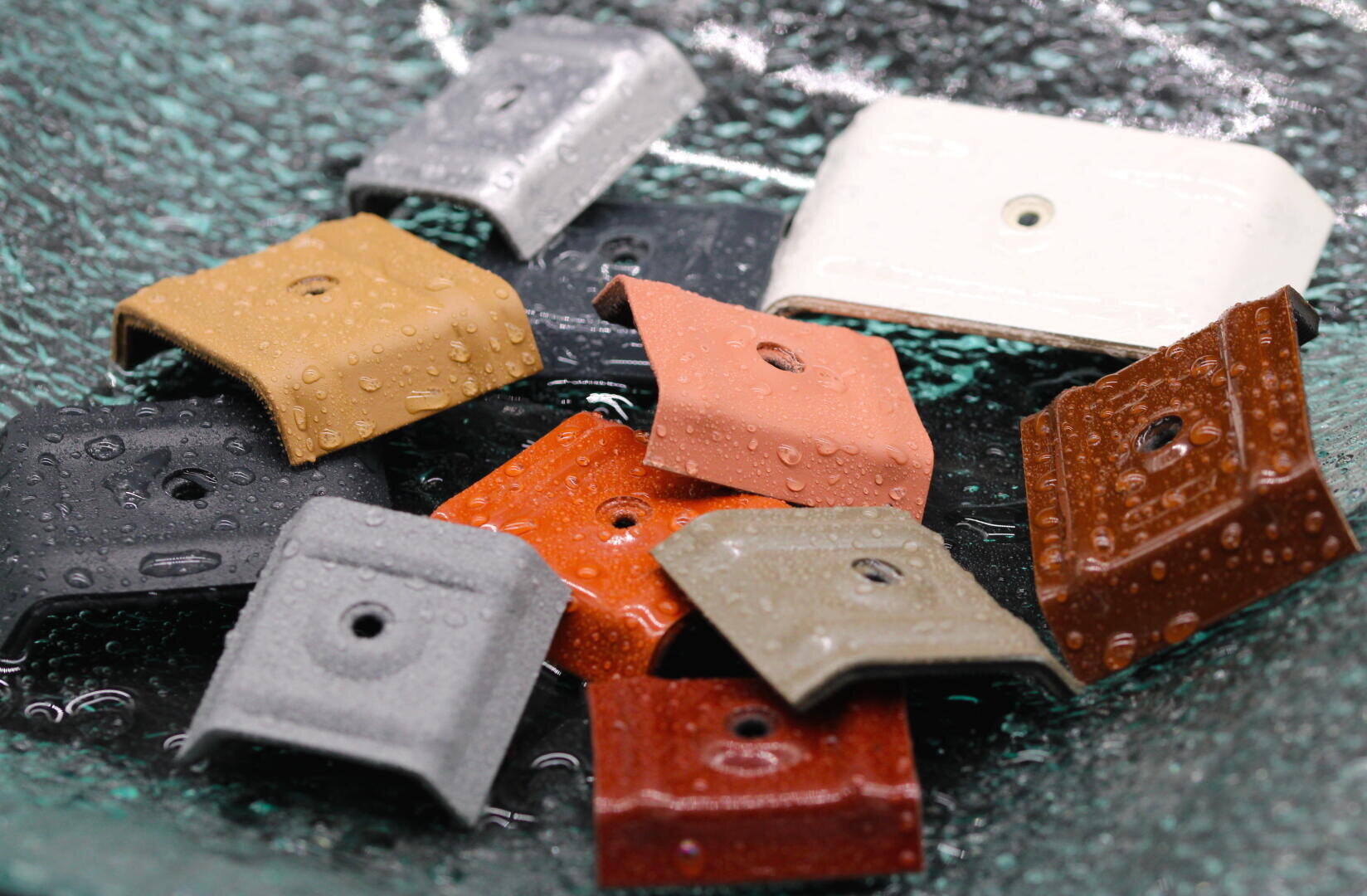 Verschiedene quadratische und rechteckige Metallfliesen in unterschiedlichen Farben, jede mit einem Loch in der Mitte und Wassertropfen auf der Oberfläche, ausgestellt auf strukturiertem Glas.