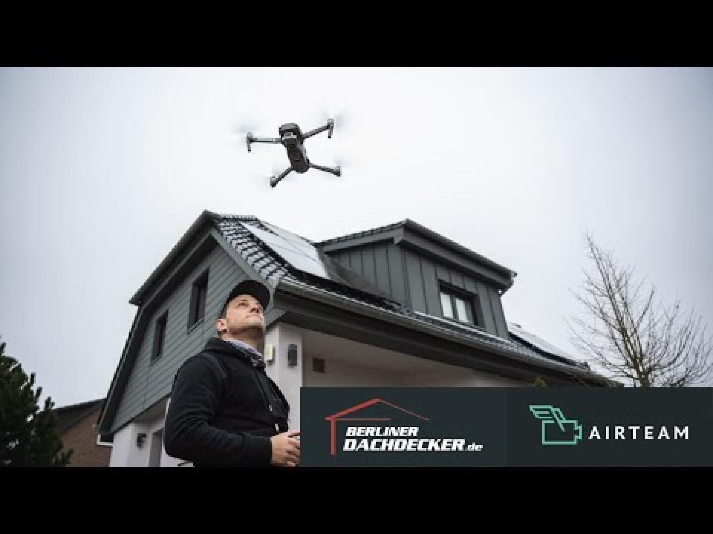 A person operates a flying drone in front of a modern house with a dark façade. The sky is cloudy. The logos of Berliner Dachdecker and Airteam can be seen in the foreground.
