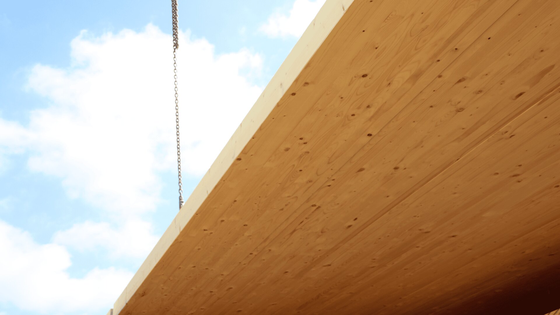 A large wooden beam or panel is being lifted by a crane against a blue sky with scattered clouds. The metal chain is attached to the wooden structure, likely during construction.