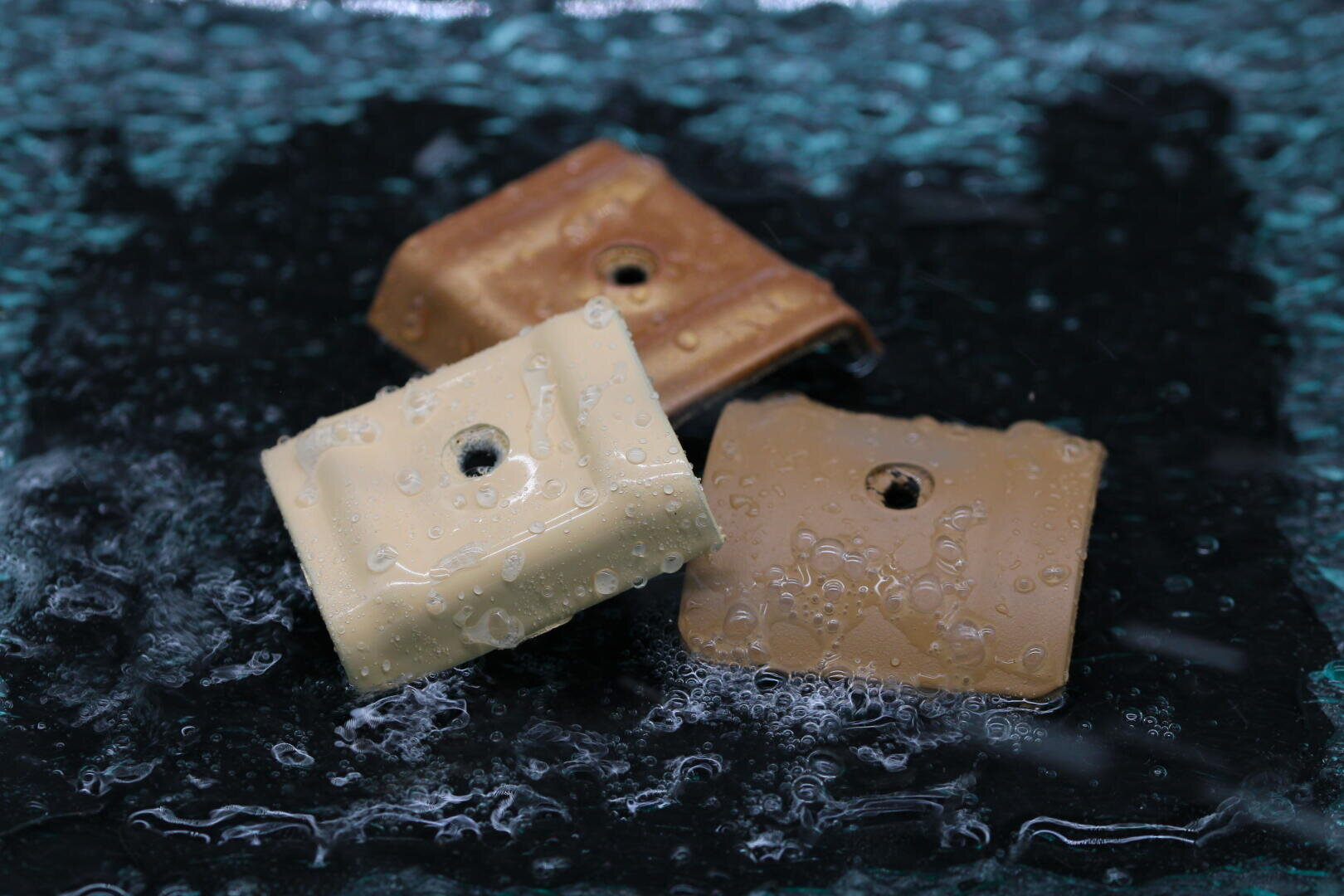 Drei rechteckige Objekte in Beige- und Brauntönen, jedes mit einem Loch in der Mitte, liegen auf einer nassen, strukturierten dunklen Oberfläche. Wassertropfen bedecken die Objekte und die Oberfläche und vermitteln ein Gefühl von Frische und Feuchtigkeit.