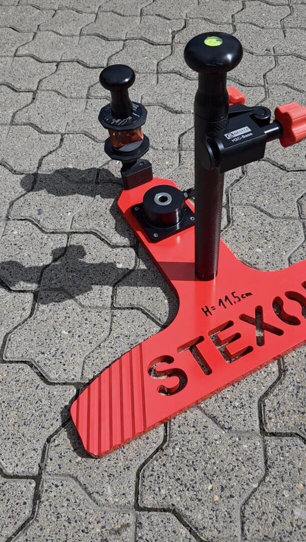 Ein rotes STEXO-Werkzeug mit schwarzen Knöpfen und einer Messskala steht auf einem grauen Pflaster mit Ziegelsteinmuster. Das Sonnenlicht wirft Schatten, und die Höhe des Werkzeugs ist mit 11,5 cm angegeben.