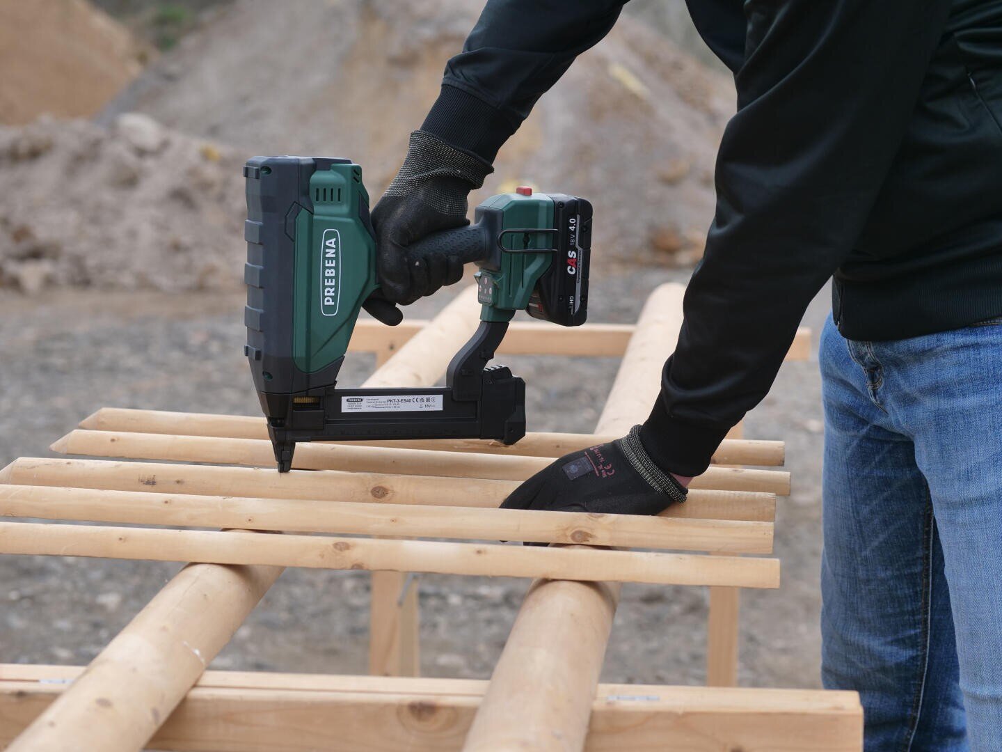 A person in black gloves uses a cordless nail gun to attach wooden slats to a wooden frame outdoors on a construction site.