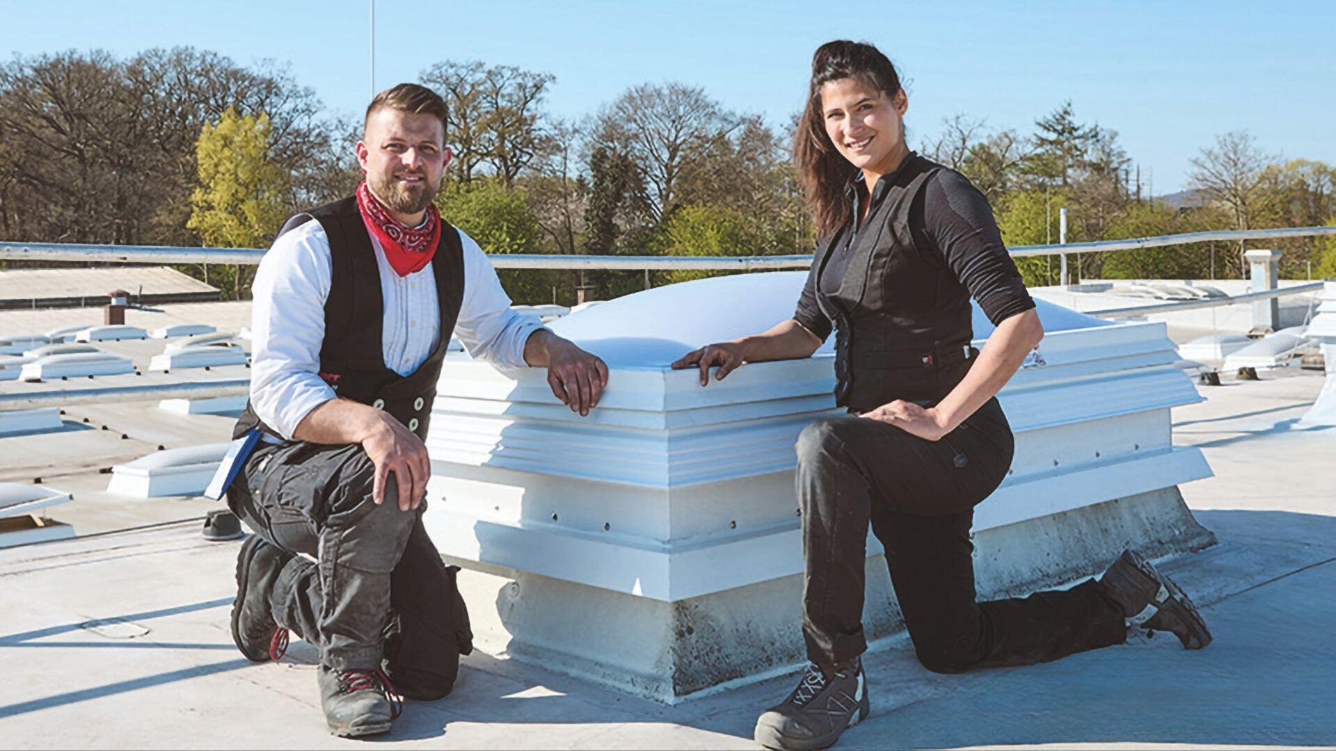 Zwei Personen in Arbeitskleidung und Stiefeln knien auf einem flachen Dach neben einer weißen Oberlichtkonstruktion, mit Bäumen und einem klaren blauen Himmel im Hintergrund.