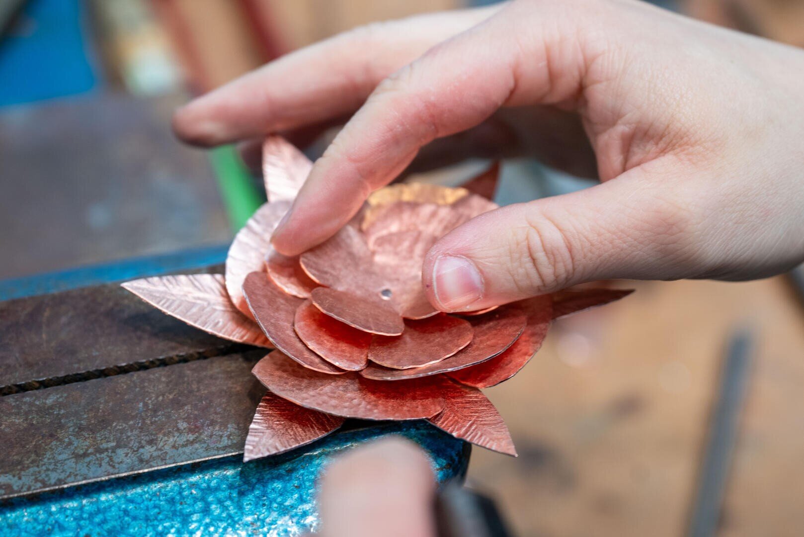 Die Hände einer Person fügen geschichtete Kupferblütenblätter in Form einer Blume zusammen, die auf einer Werkbank bei der Metallbearbeitung oder der Herstellung von Schmuck festgehalten werden.