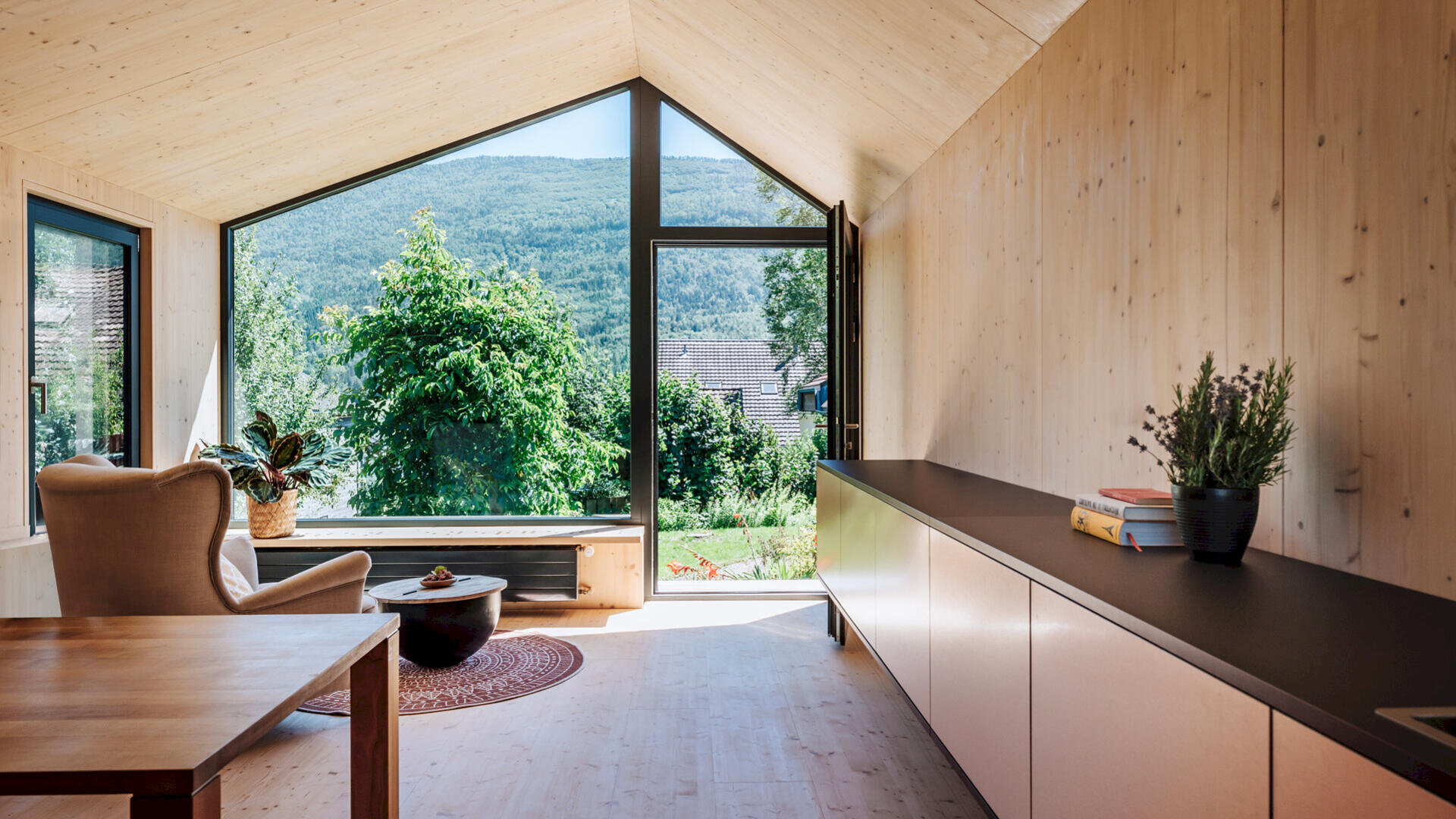 Ein modernes, minimalistisches Wohnzimmer mit heller Holzvertäfelung, großen raumhohen Fenstern mit Blick auf grüne Bäume und Berge, einem Sessel, Topfpflanzen und einem niedrigen Schrank mit Büchern und einer Vase.