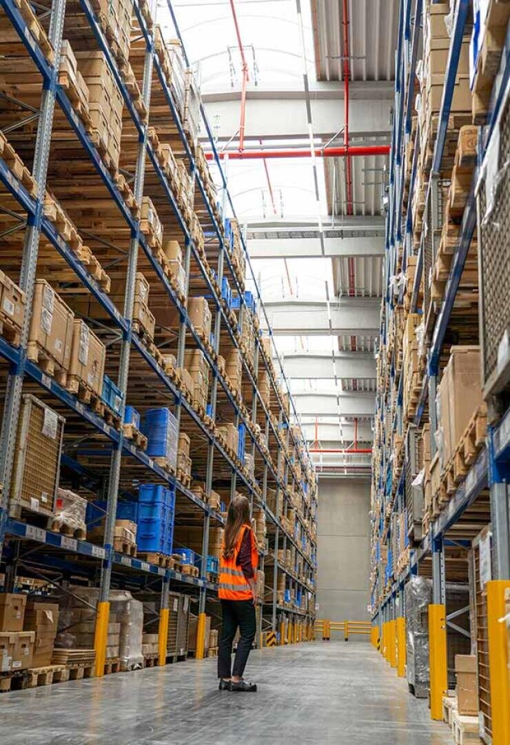 Eine Frau, die eine Warnweste trägt, steht in einem großen Lagergang, umgeben von hohen Regalen mit Kisten und Paketen unter einer hellen, lichtdurchfluteten Decke.
