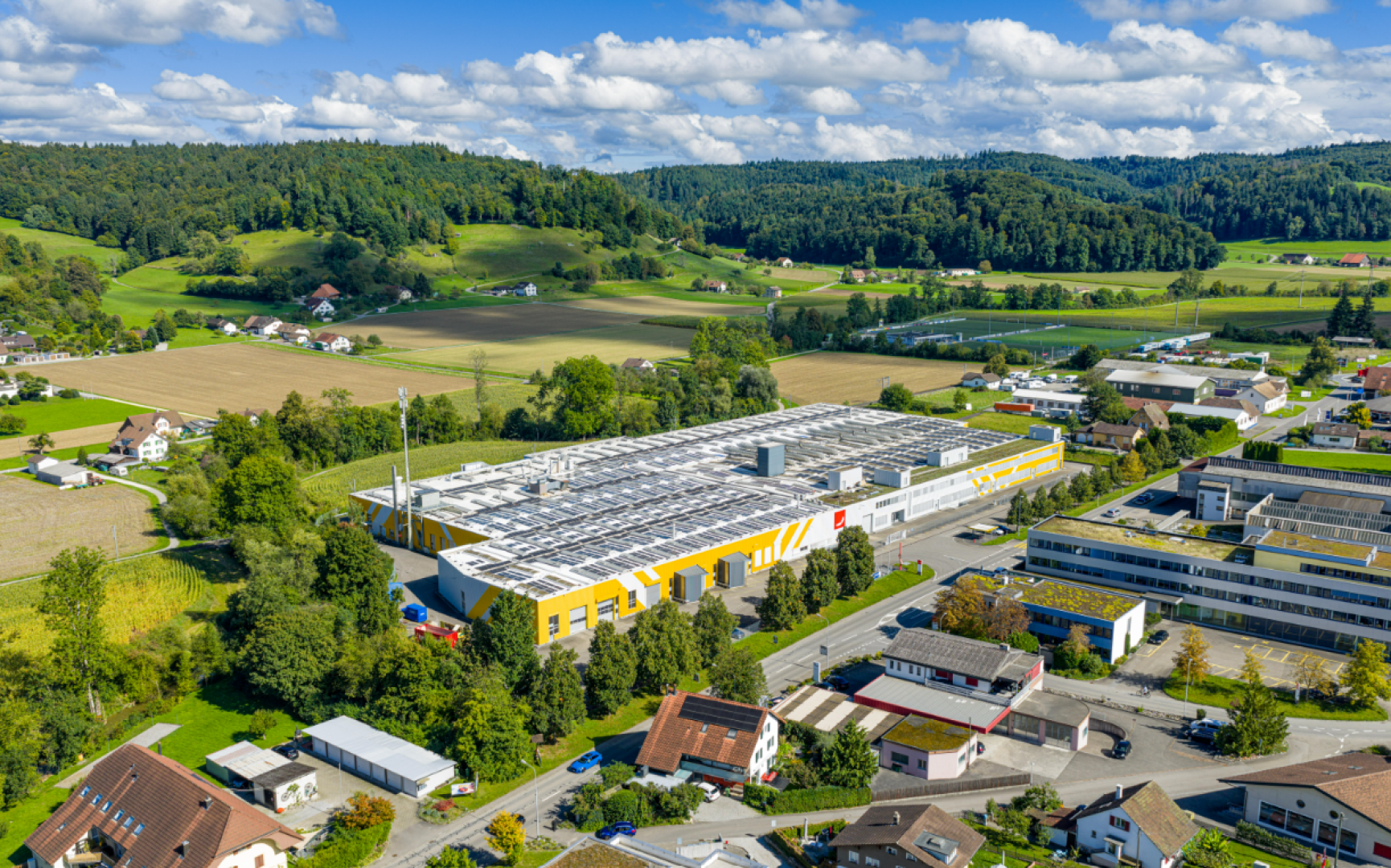 Aerial view of a large yellow and white industrial building with solar panels on the roof, surrounded by smaller buildings, fields, trees and hills under a partly cloudy sky.
