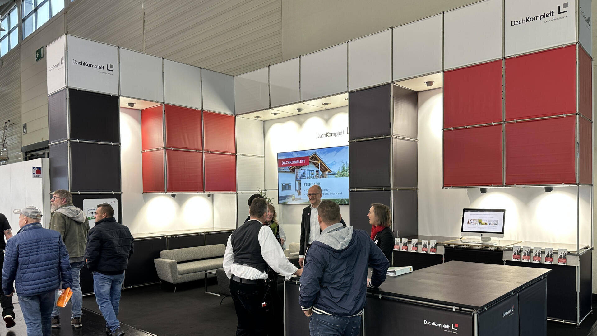 Ein moderner Messestand mit roten und weißen Paneelen zeigt das Logo von DachKomplett. Mehrere Personen unterhalten sich auf dem Stand, der mit Broschüren, einem Monitor und beleuchteten Regalen ausgestattet ist.
