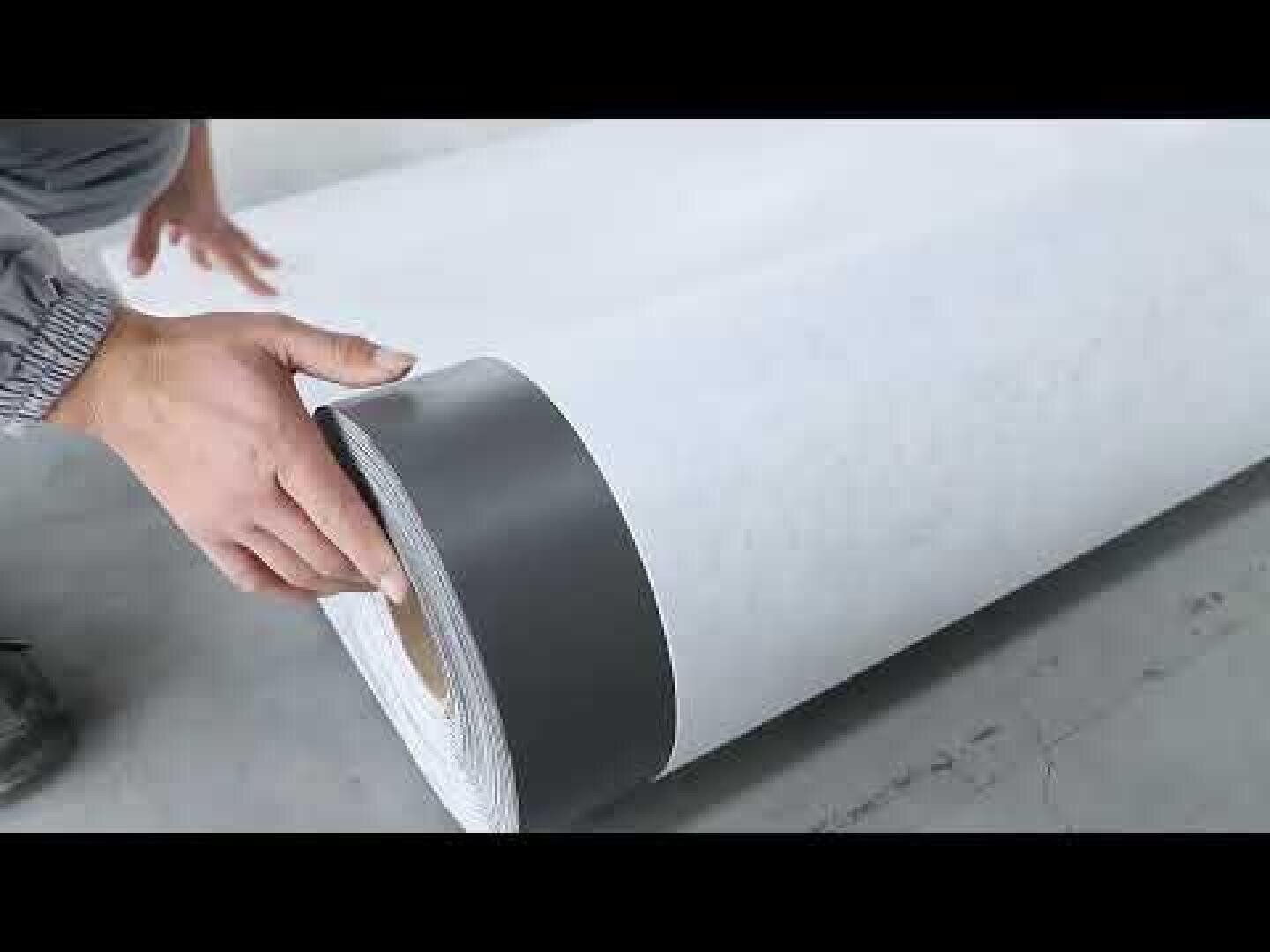 A person's hands unroll a large roll of white fabric with a gray border strip that lies on a smooth, light gray floor.