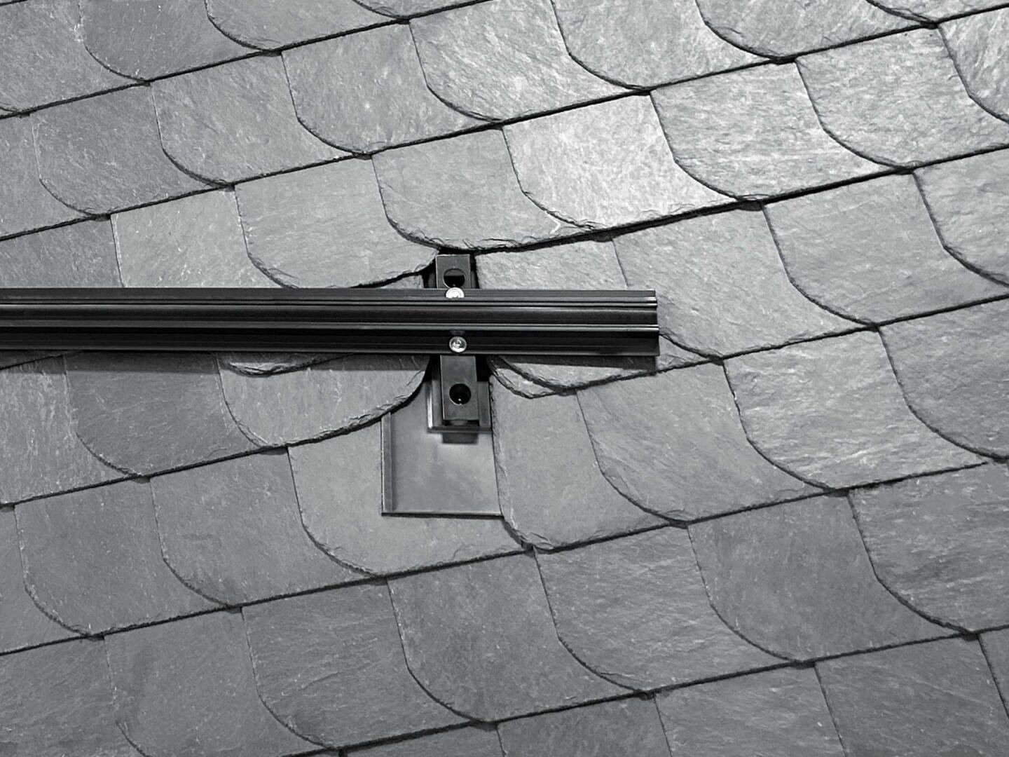 Auf einem mit überlappenden grauen Schieferziegeln gedeckten Dach sind eine Metallhalterung und eine Schiene installiert, die wahrscheinlich zur Befestigung von Sonnenkollektoren oder ähnlichen Geräten dienen.