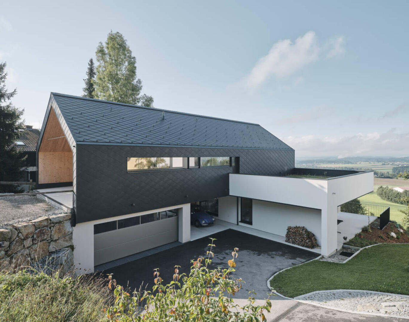 Ein modernes Haus mit schwarzem Schrägdach, weißen und dunklen geometrischen Außenwänden, einer großen Garage und einer Einfahrt, das auf einem begrünten Hügel mit offenem Himmel im Hintergrund steht.