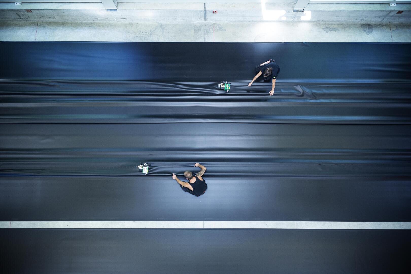 Zwei Personen arbeiten in einer Industrieumgebung an großen, glatten schwarzen Platten, wobei sie mit Handgeräten das Material pressen oder glätten; die Szene ist von oben zu sehen.
