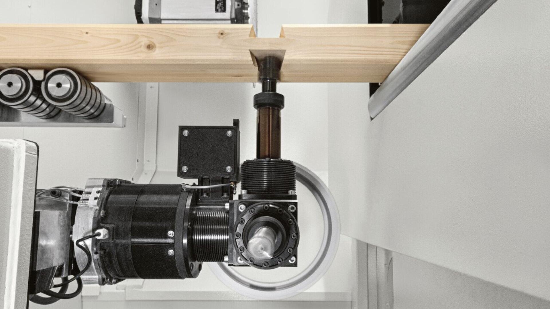 A robotic arm equipped with a rotating tool is precisely cutting or shaping a wooden beam inside an industrial setting, with machinery components visible around the workspace.